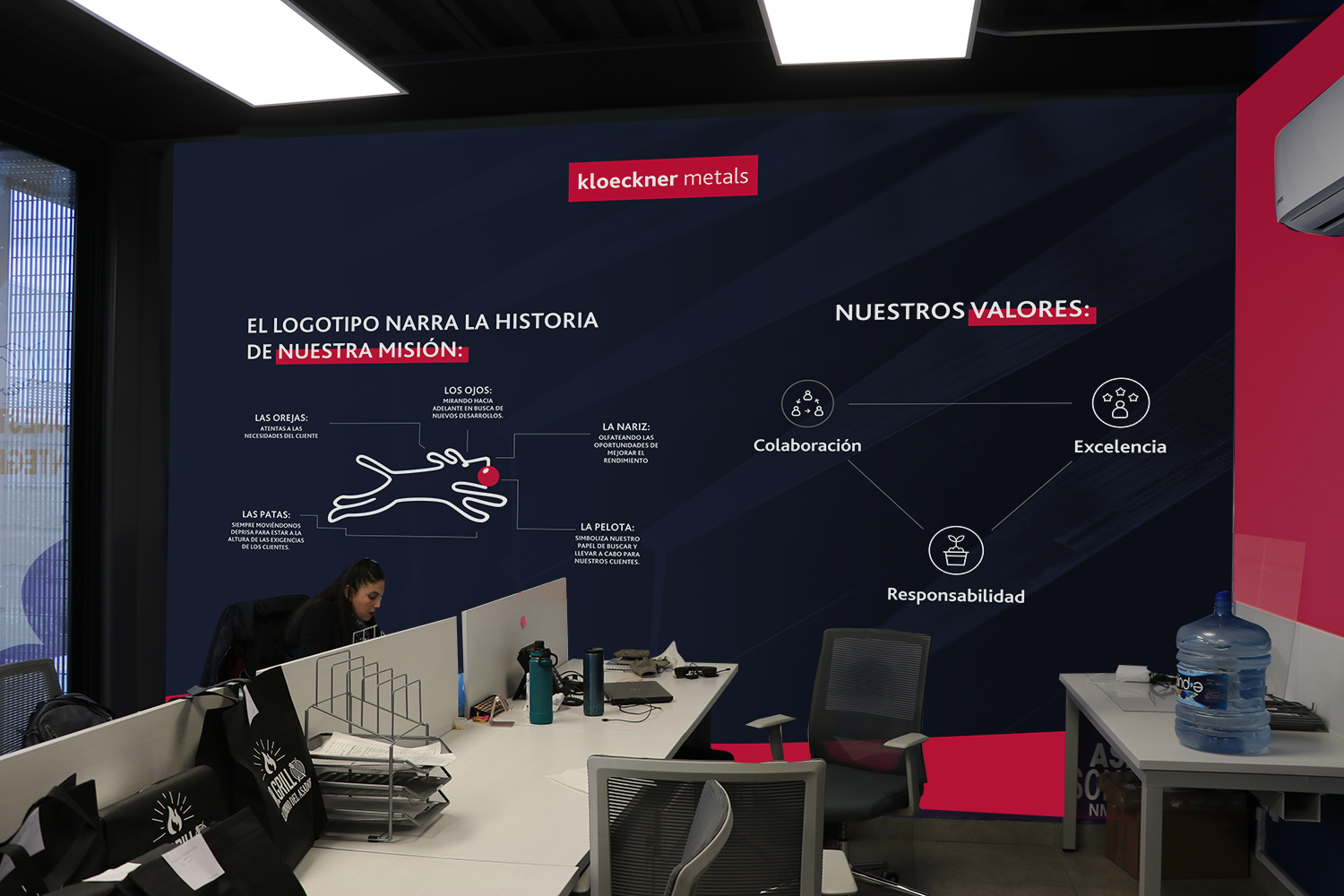 Office space with a large dark blue wall displaying company logo and mission statement in Spanish, including diagrams and words such as 'Colaboración', 'Excelencia', and 'Responsabilidad'. A woman works at a desk in the foreground, with office suppli