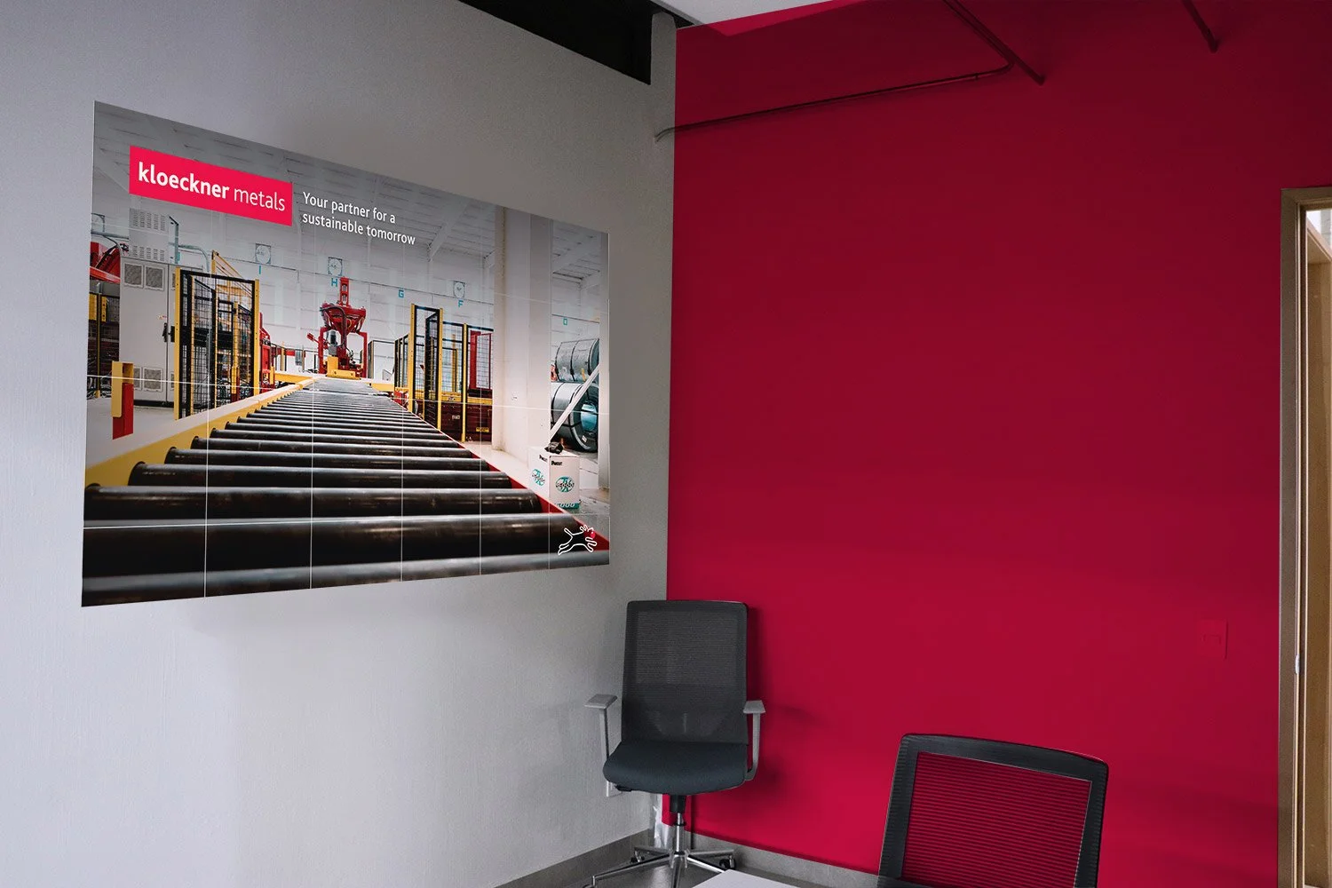 A waiting room with a red accent wall, a framed poster of an industrial metal manufacturing facility, a black office chair, and a partial view of a black mesh chair.