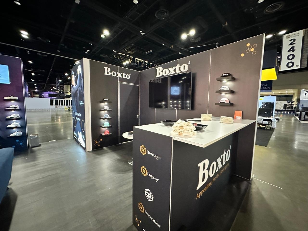 Exhibition booth for Boxto showing hats on wall displays and a table with folded cloths. The booth features a black and gray color scheme with company branding and a large screen.