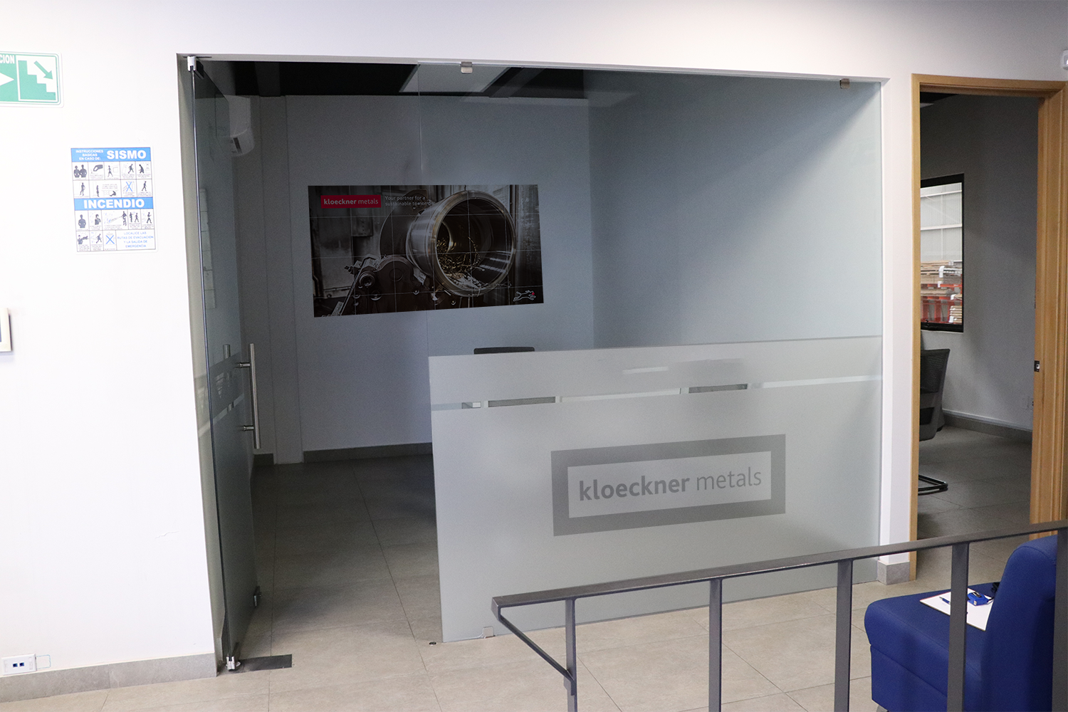 Office reception area with a glass wall displaying 'kloeckner metals' logo, a poster of a turbine engine, a blue chair with a notepad, and informational signs on the wall.