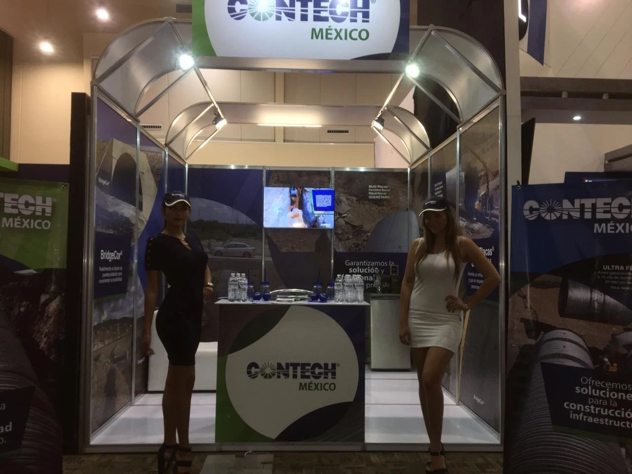 Trade show booth with two women standing inside, branding for CONTECH MEXICO, advertising construction and infrastructure solutions, illuminated display with bottles of water on counter, promotional banners on sides, digital screen showing constructi