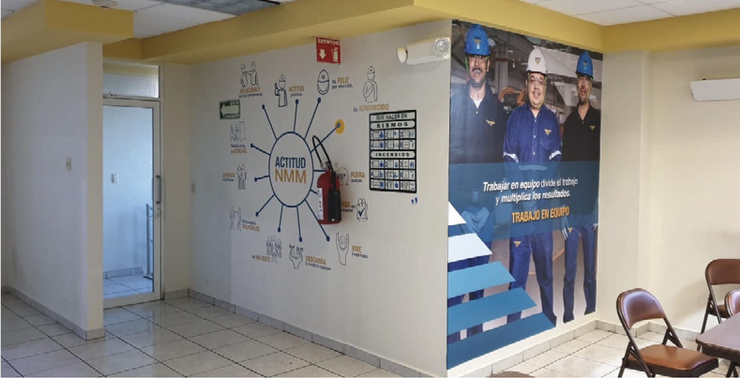Interior of a building with a wall featuring safety and teamwork posters, including a circular diagram titled "Actitud NMM," and a large poster of three workers wearing safety helmets with a motivational message, along with chairs and a doorway.