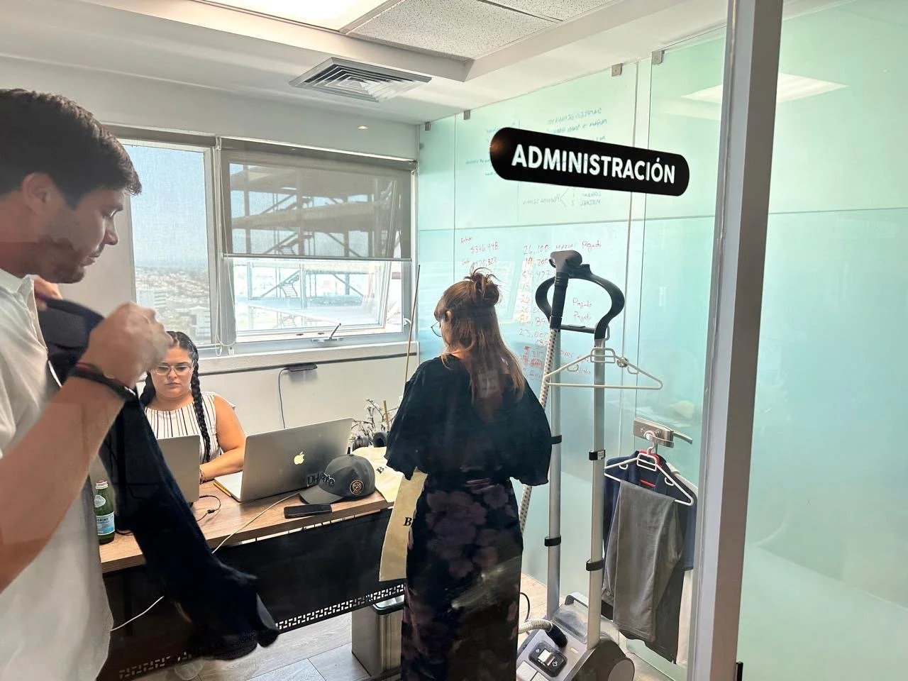 Office scene with three people, one man and two women, inside a glass-walled room labeled 'Administración'. Woman in black appears to be using a treadmill. Man is looking at his jacket, and woman with glasses and a striped top is working on a laptop 