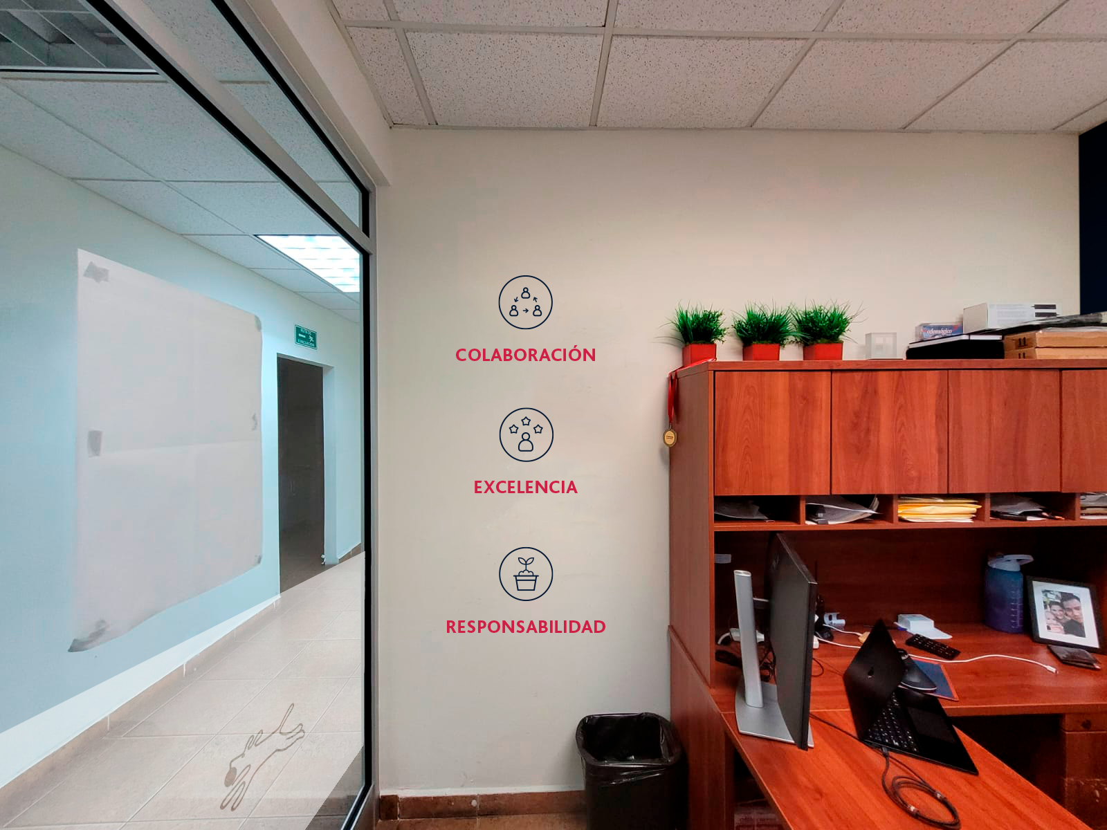 An office corner with a wooden desk with computer, plants, and files; on the white wall, there are three icons with words: collaboration, excellence, responsibility.