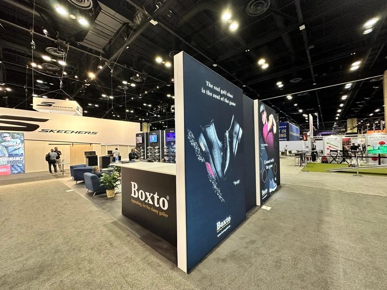 A trade show booth for the brand Boxto displaying golf shoes, with promotional signage and a seating area, located near the Skechers section.