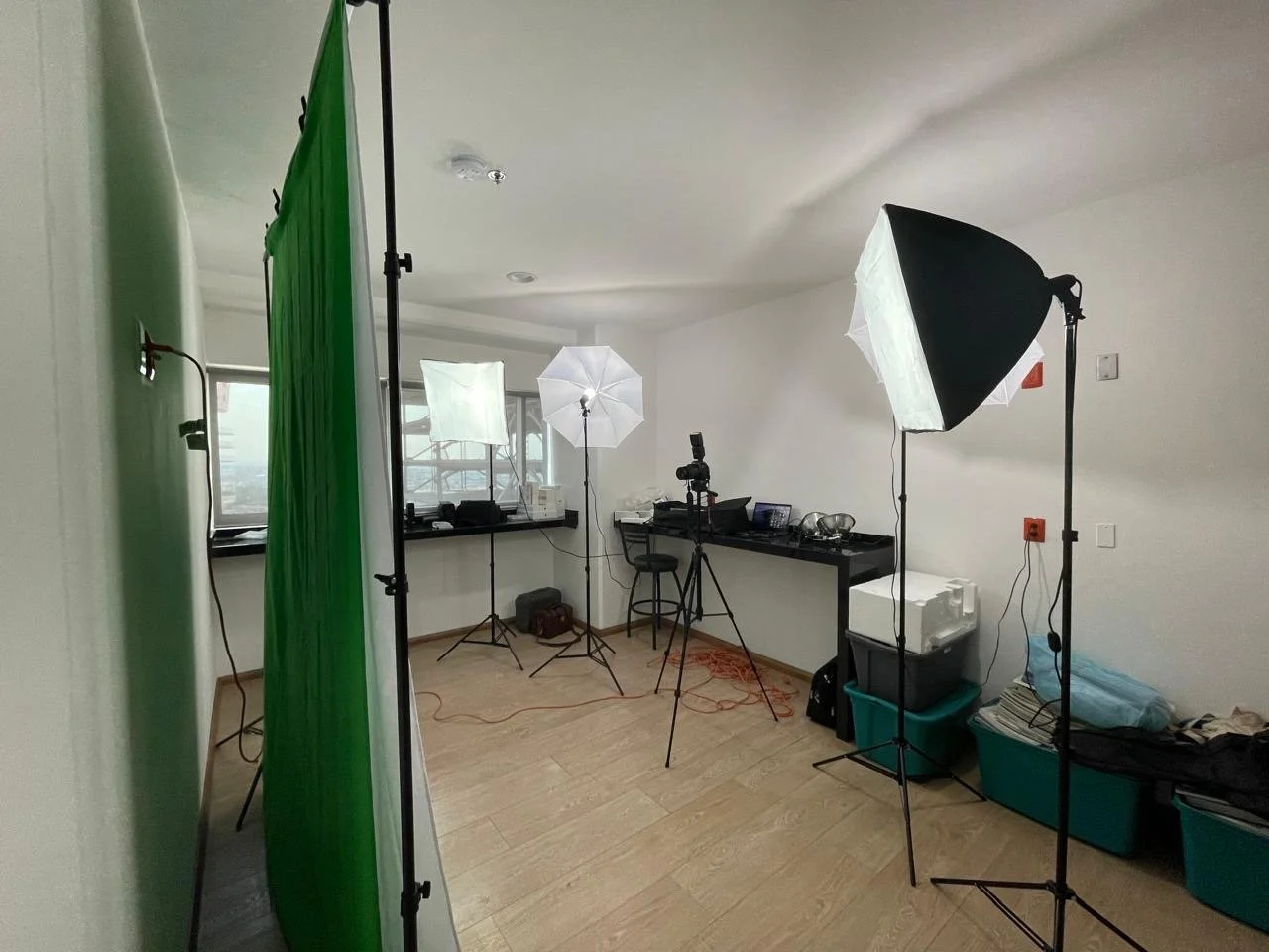 Indoor makeshift photography studio with green screen, softbox lights, tripods, and various equipment.
