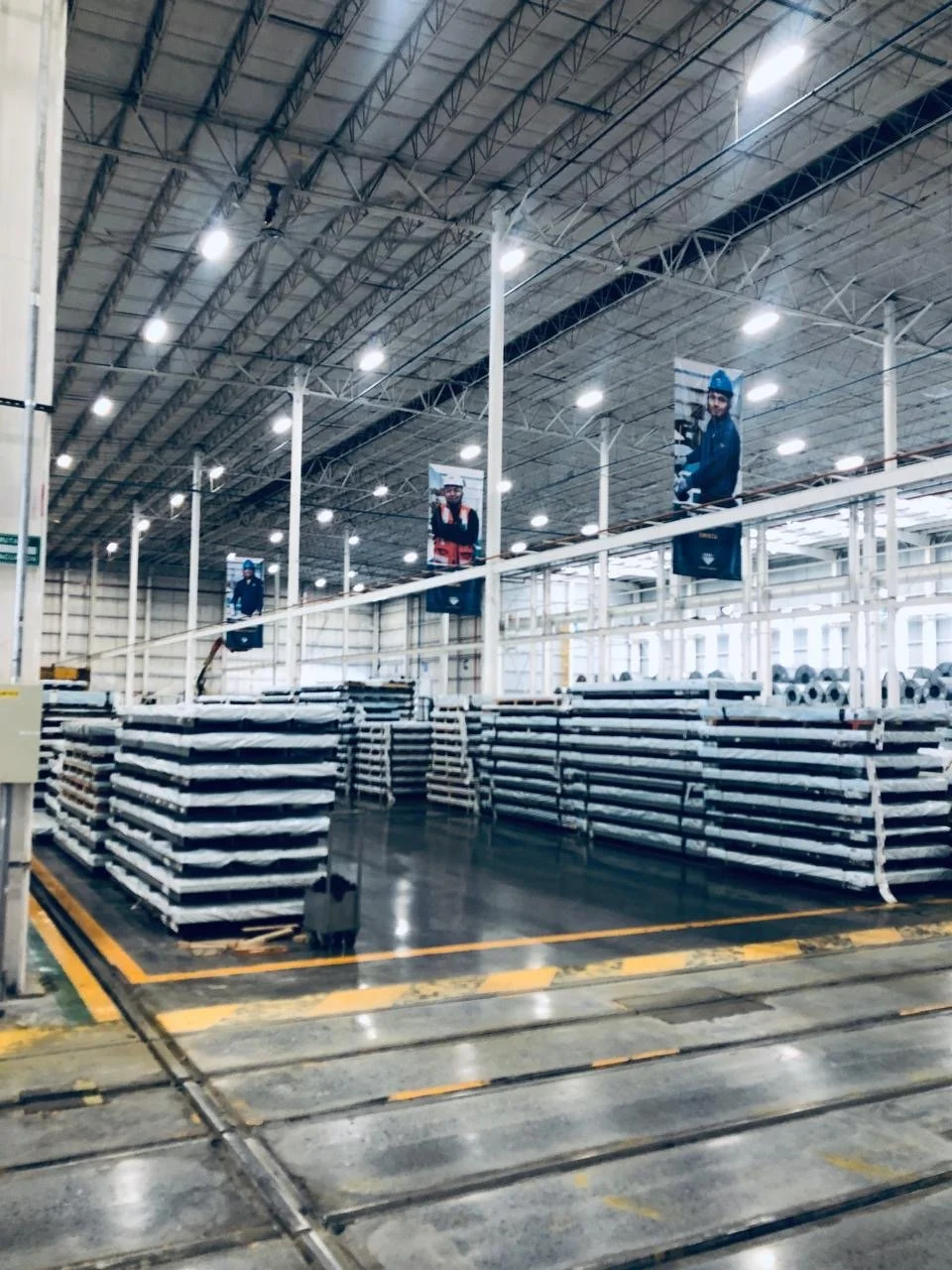 Interior of a warehouse or factory with stacks of metal pipes, safety rails, and industrial lighting fixtures on the ceiling.