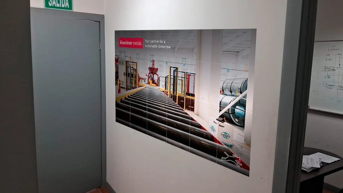 Interior of an industrial facility with a large pipe conveyor system in a hallway, advertisement poster for Kloeckner Metals on the wall, and a door with a green exit sign.