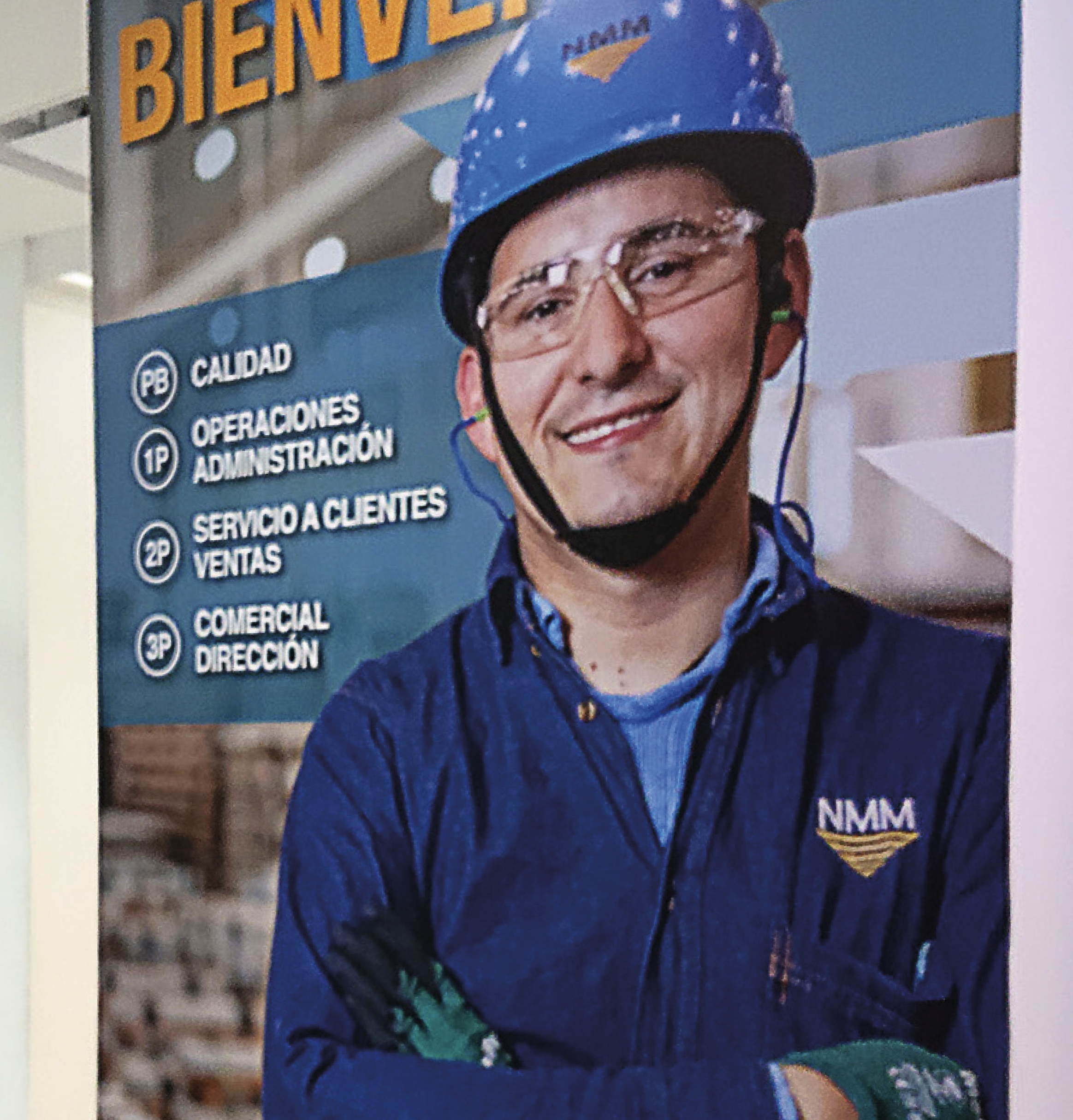A man in safety gear, including a blue helmet and glasses, standing in front of a sign with company name NMM and a list of departments in Spanish, such as quality, operations, customer service, and sales.