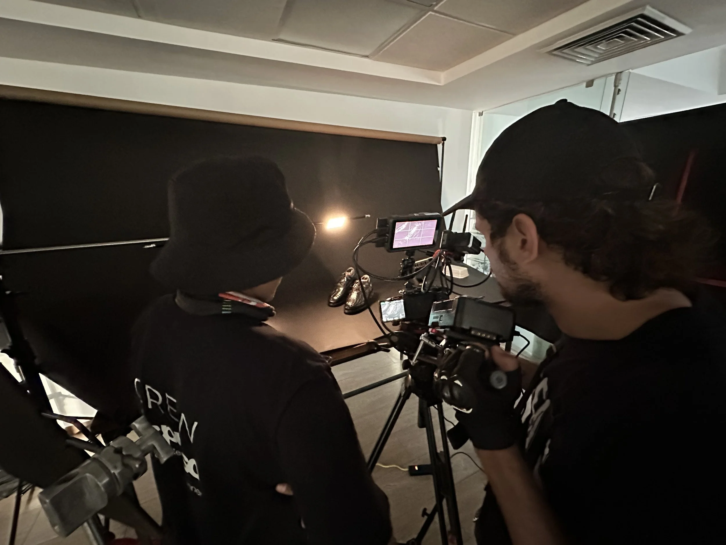 Two people working with a camera and monitor on a film set, one person operating the equipment and the other person observing, in a dark room with a monitor screen and equipment.