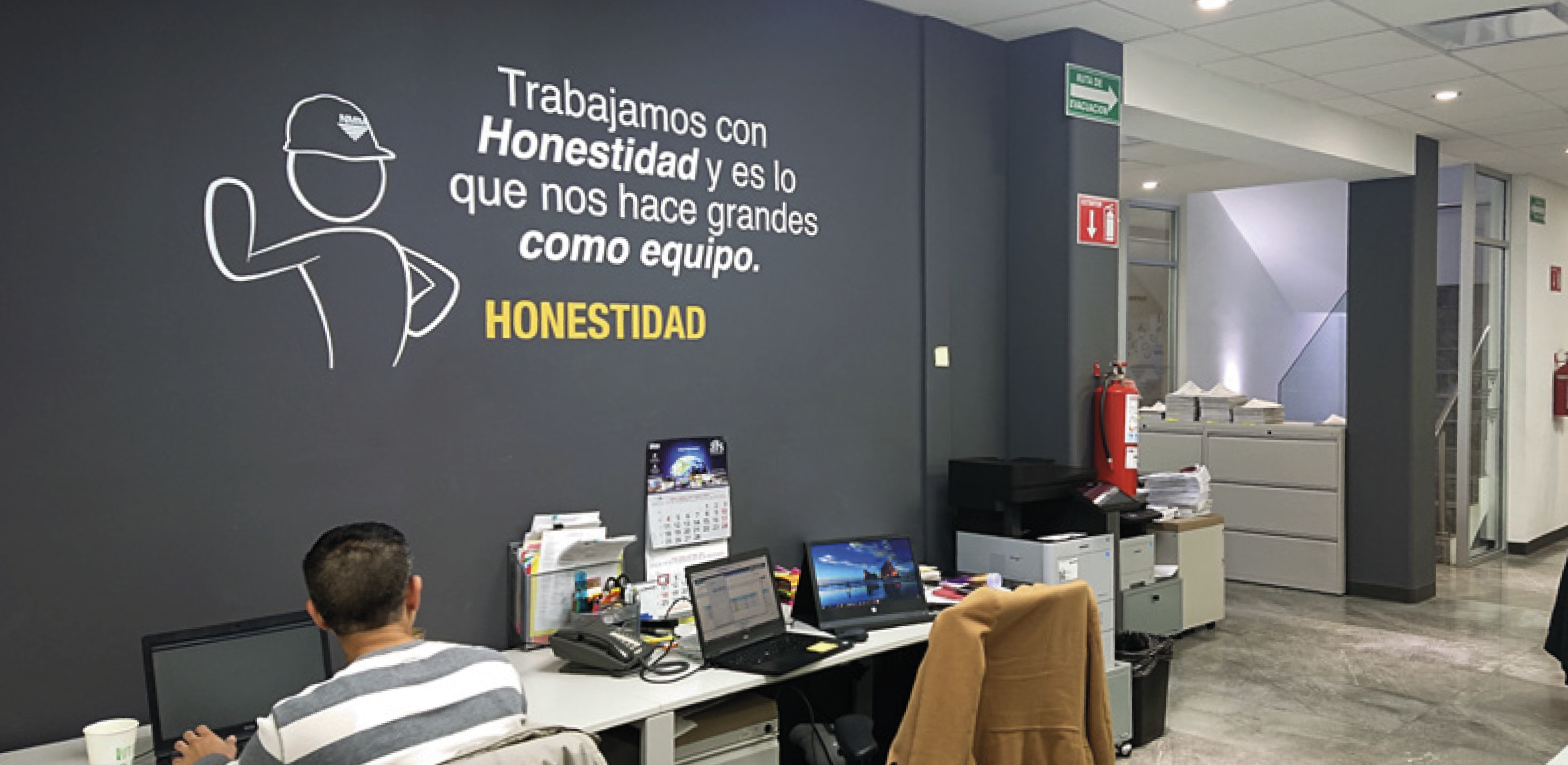 Office space with a dark gray wall featuring a quote about honesty and teamwork, a desk with laptops, a printer, and paperwork, and a man working at the desk.