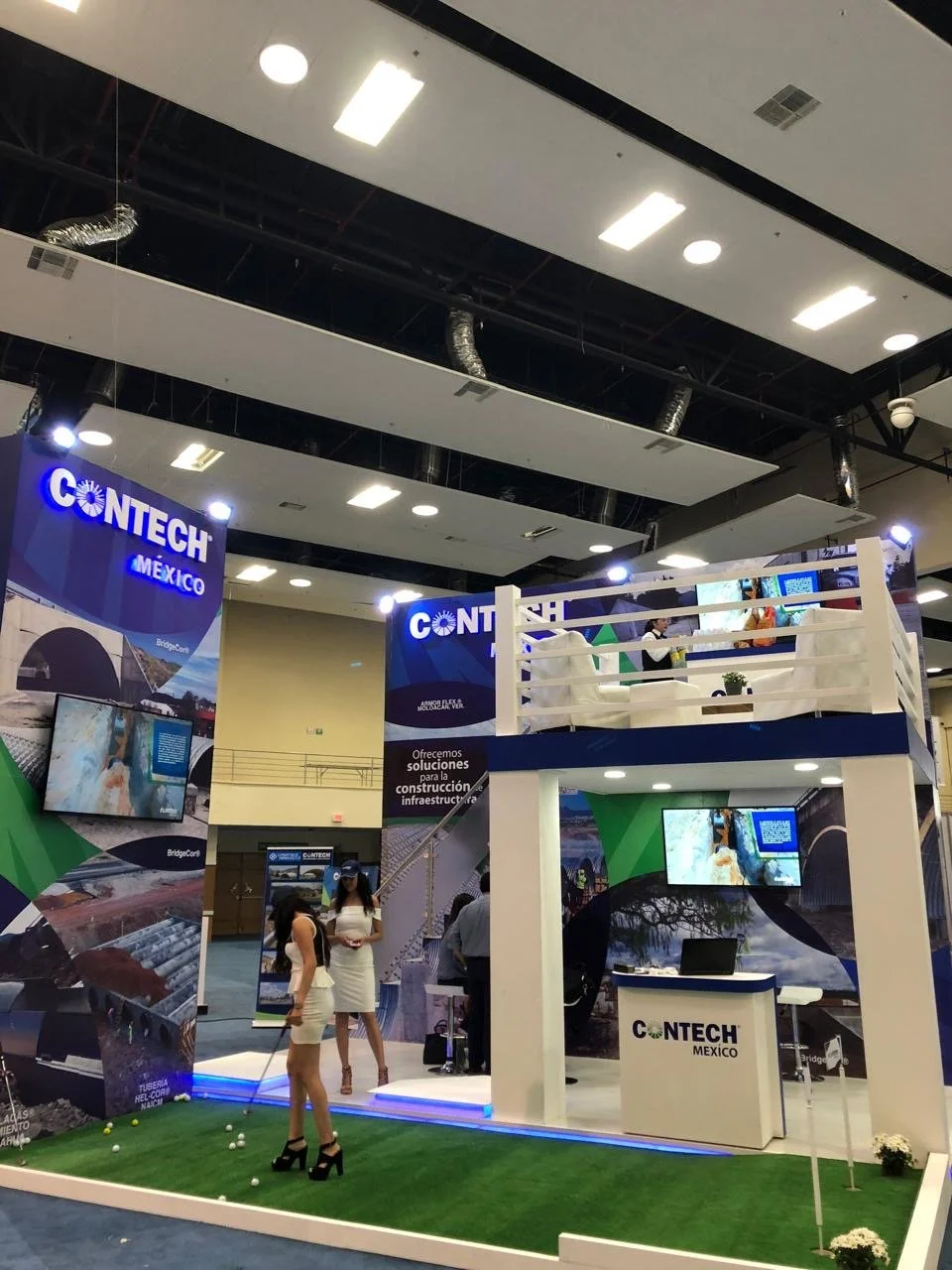 Trade show booth for CONTECH Mexico with displays, monitors, and a small green area with two women dressed in white and black overseeing activities.