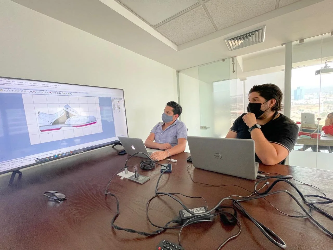 Two men wearing face masks sit at a conference table, viewing a large monitor displaying a digital design of a sneaker, with laptops and cables in front of them.