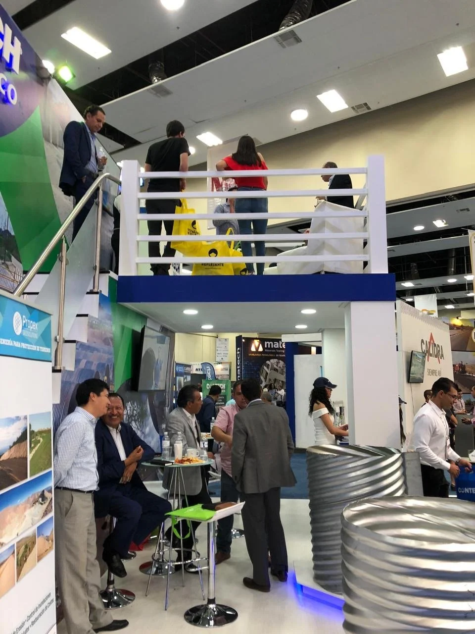 People attending a trade show with a two-story booth, some on the upper level and others conversing on the lower level among booths with promotional materials and displays.