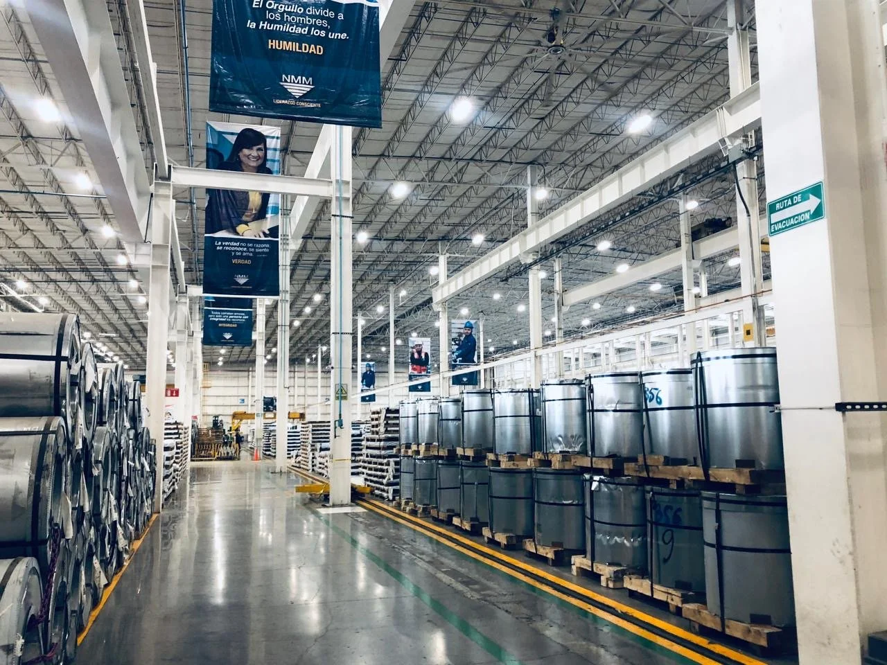 View inside a large industrial warehouse or factory with metal coils stacked on the left and large metal containers on the right, with a worker in blue uniform and helmet on the right. Overhead lights illuminate the space with high ceiling and struct