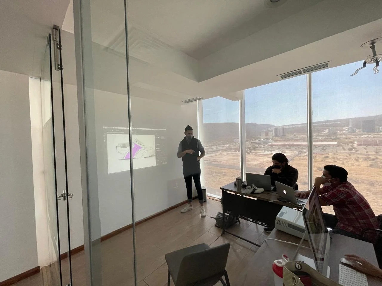 A person is giving a presentation in a conference room with large windows, two other people are seated at a desk with laptops, and a projection screen at the front displays a sneaker illustration.