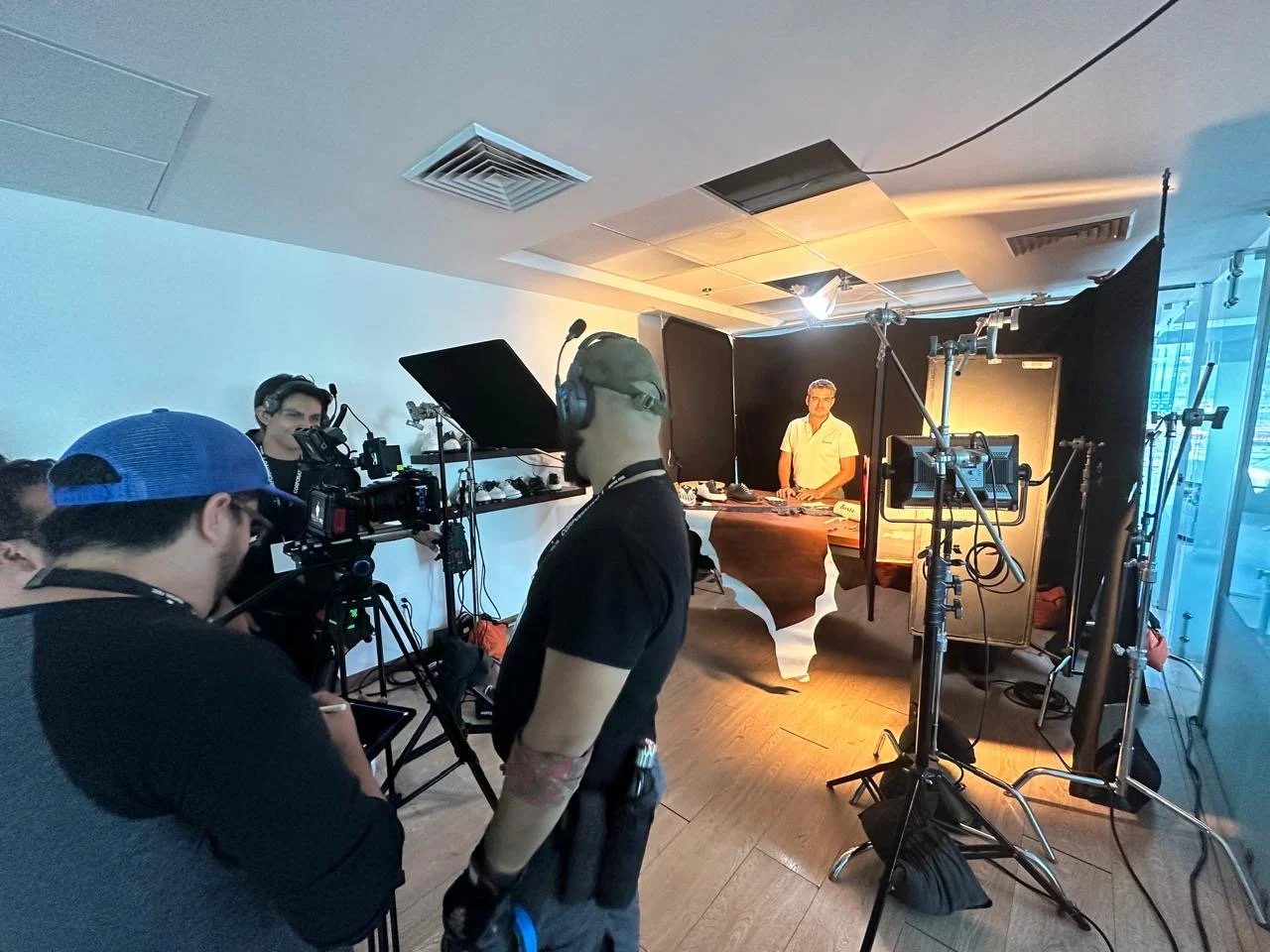Film crew setting up for a shoot with a man at a desk, various lighting and camera equipment around, inside a modern room with a window.