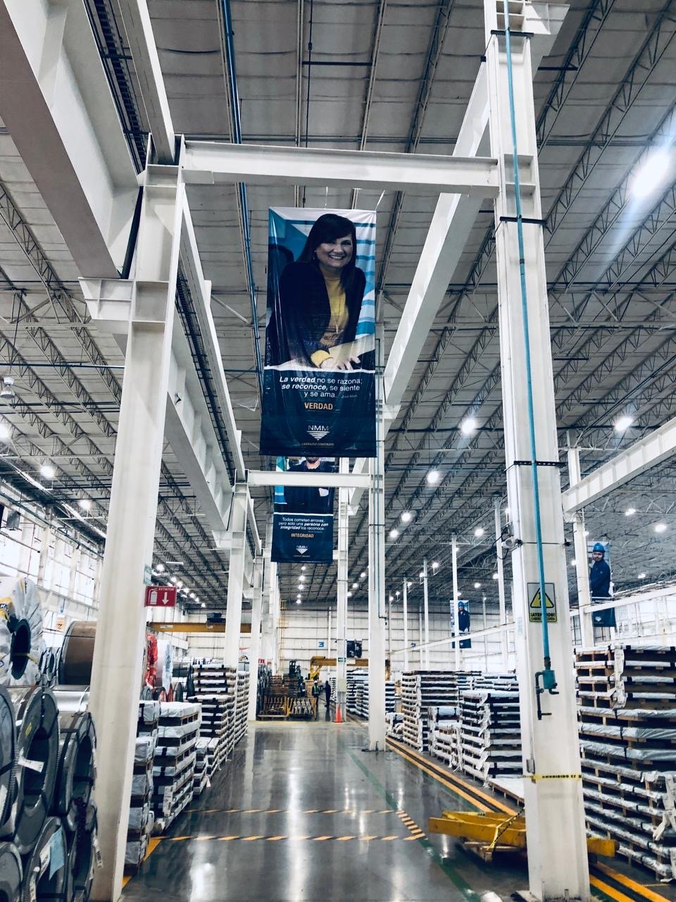 A large industrial warehouse with high ceilings, metal beams, and bright lighting. Two banners hang from the ceiling displaying a smiling woman with dark hair, dark blazer, and yellow shirt, along with Spanish text. Metal shelves stocked with various
