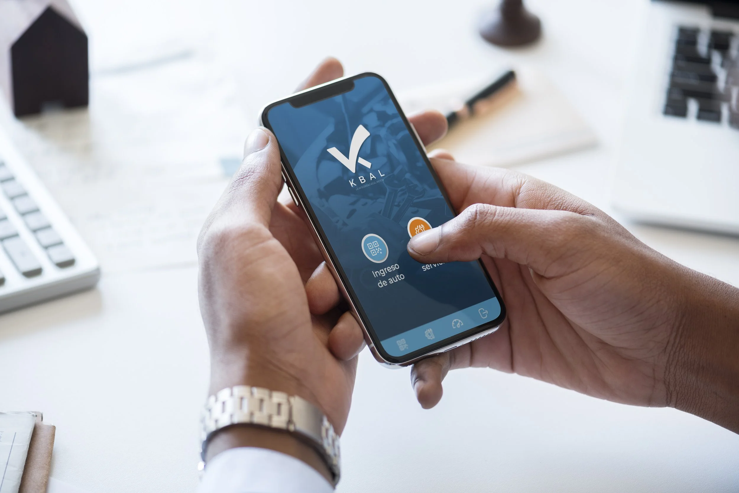 Person holding a smartphone displaying an app interface with the logo 'V KBAL' and options for car entry and service, on a white desk with a keyboard, notepad, and office supplies.