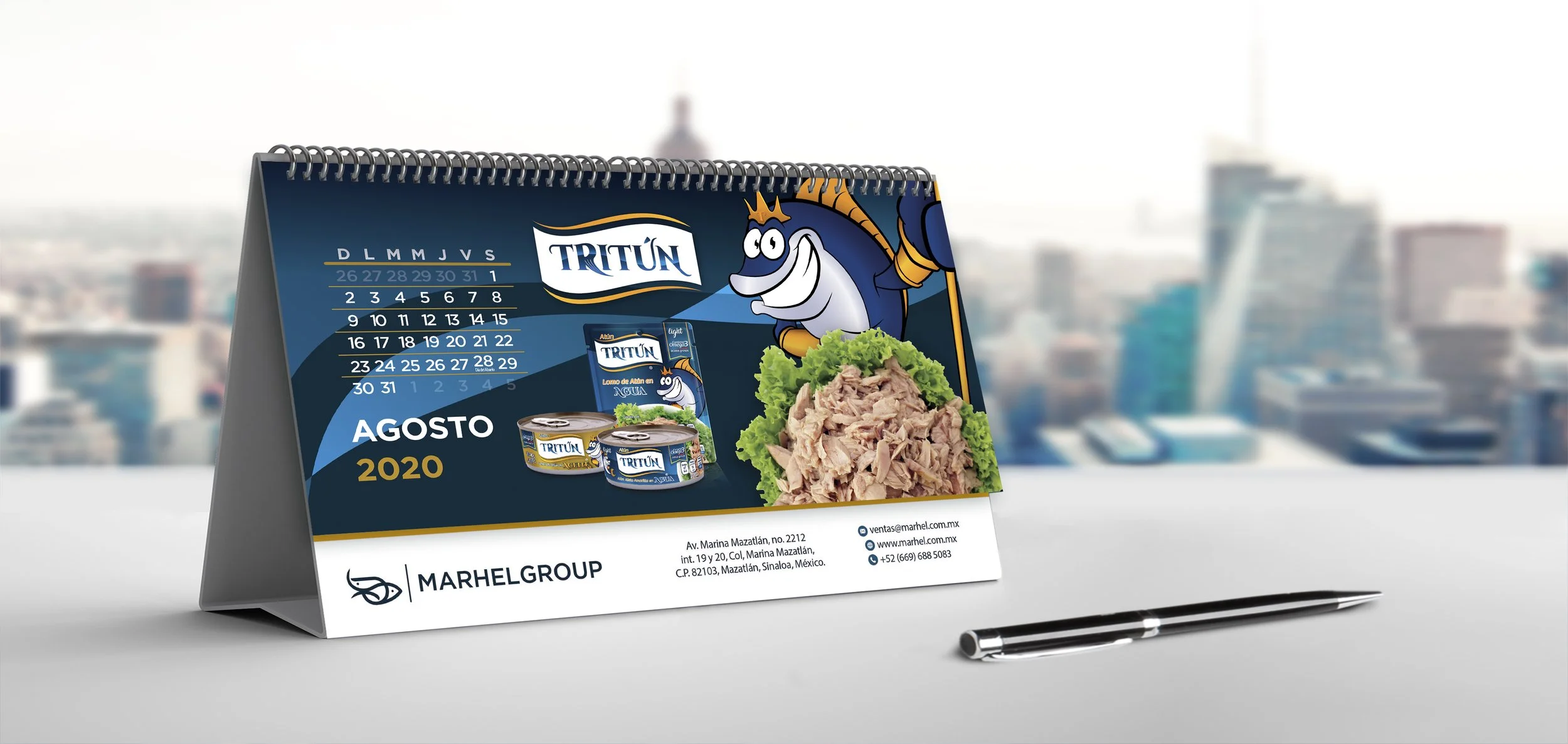 Desk calendar with August 2020 date, featuring Tuna brand canned tuna, a cartoon tuna mascot, and a salad, with a pen on a white surface, blurred city skyline in the background.