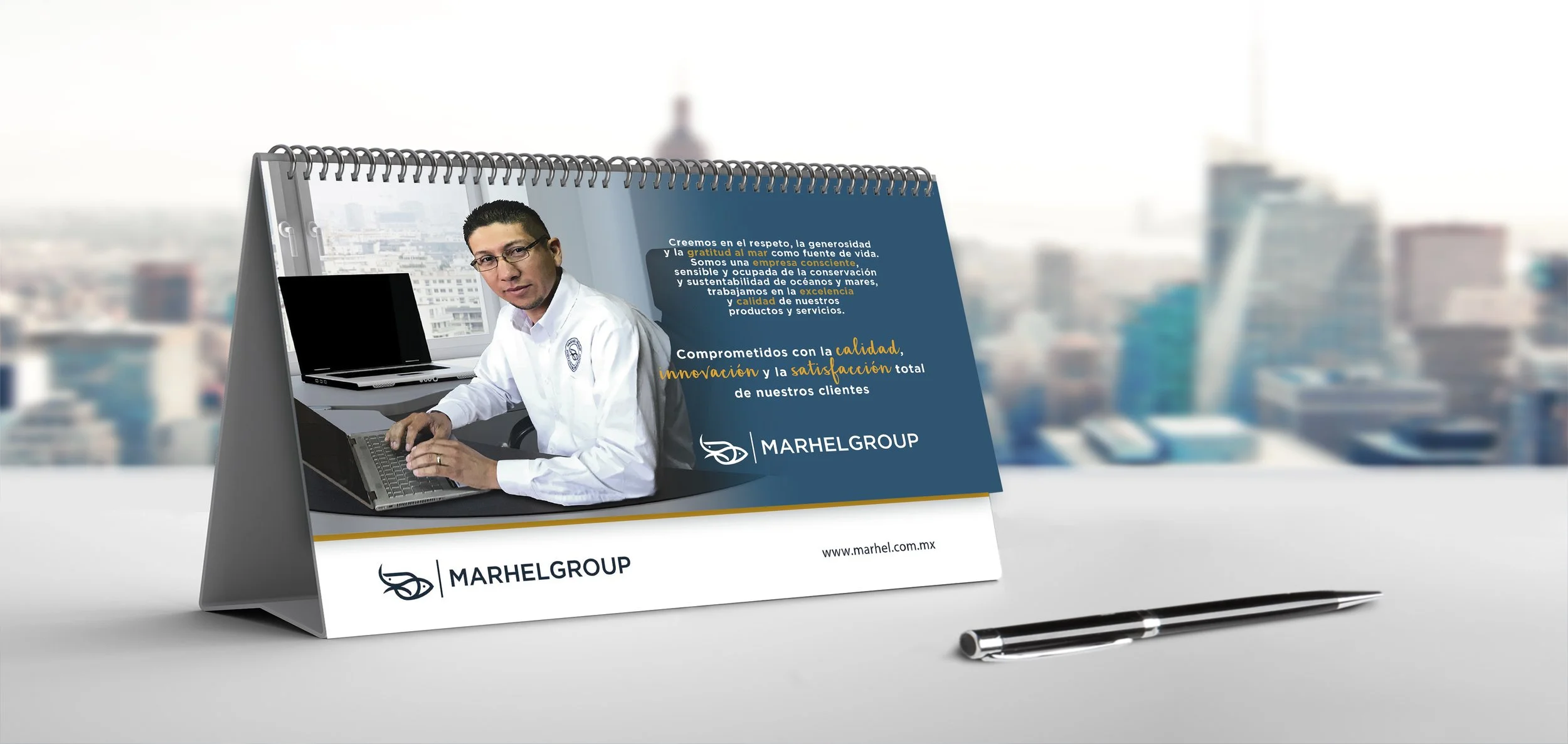 A desk calendar with the MARHEL GROUP logo, featuring a photo of a man in a white shirt sitting at a desk with a laptop. The calendar has a city skyline in the background and contains text in Spanish about the company's values and commitment to quali