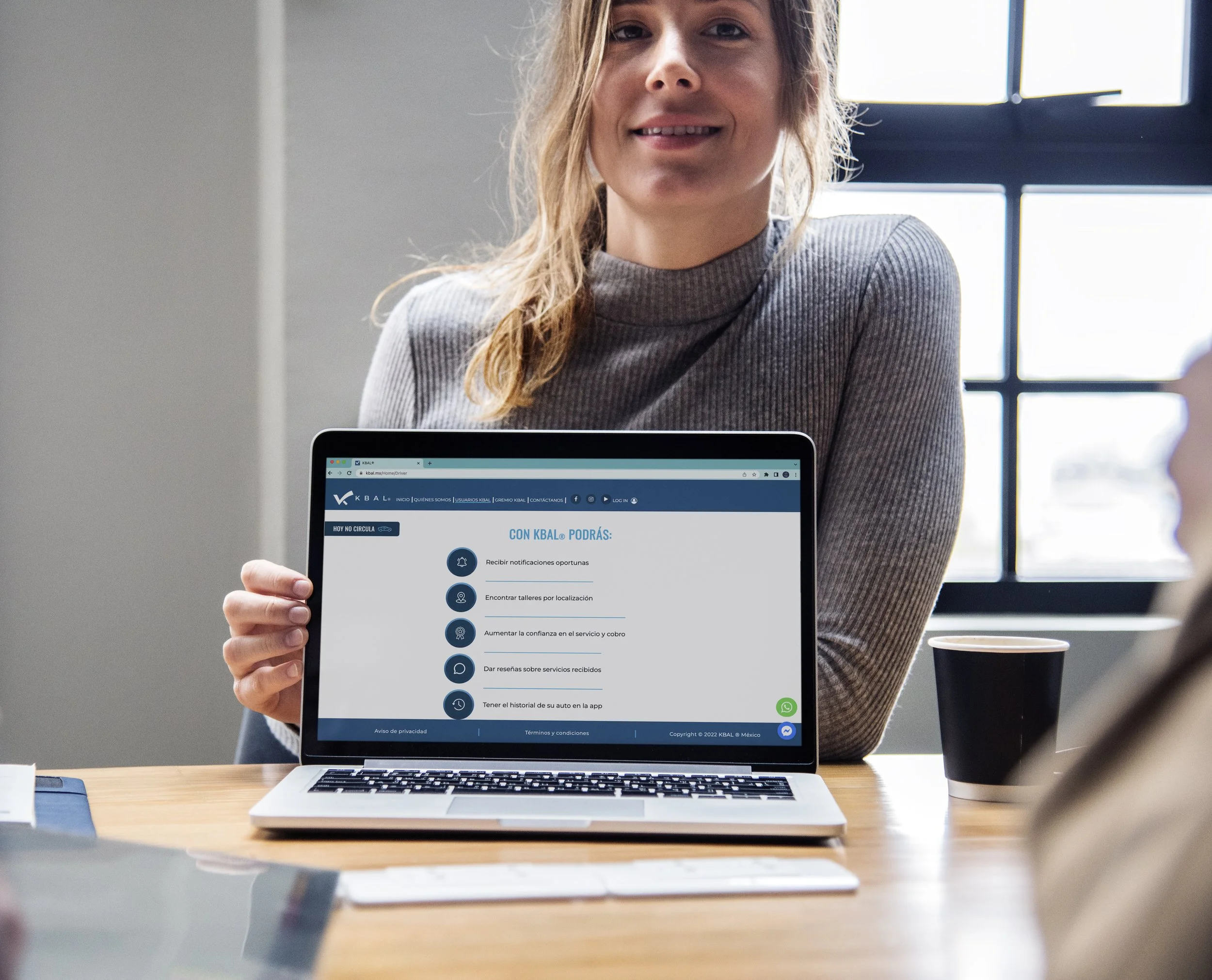 A woman holding a laptop displaying a website in Spanish, with a coffee cup on a wooden table, sitting near a window.