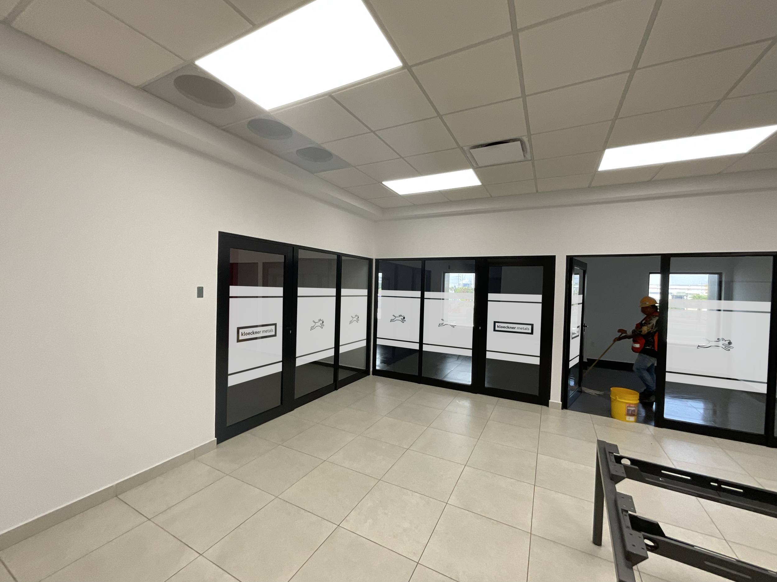 Interior of an office space with glass partition walls, a man in a hard hat cleaning the floor, and construction tools and supplies present.