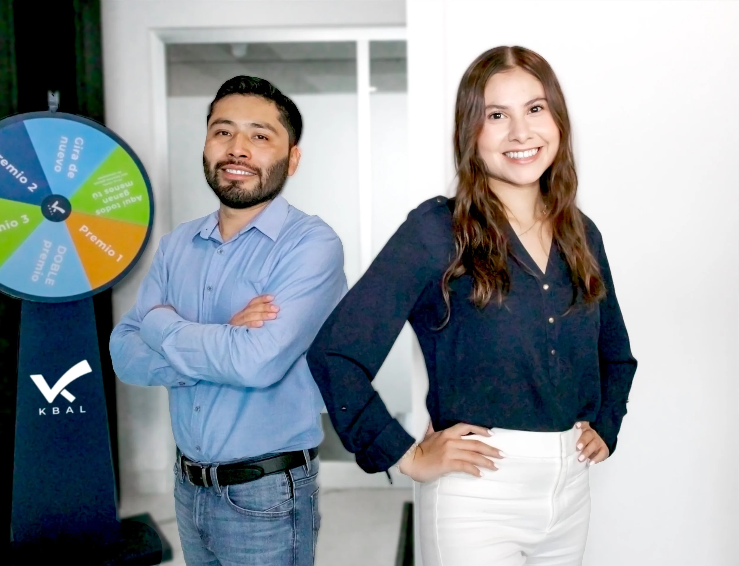 Two people, a man in a blue shirt with arms crossed and a woman in a dark blue blouse with one hand on her hip, standing indoors near a prize wheel with a white background.