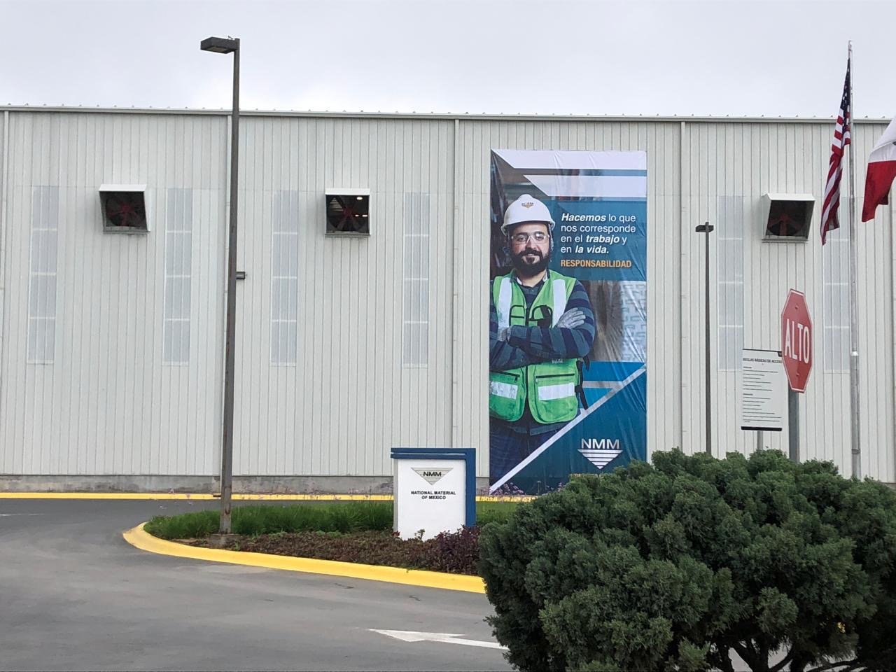 Large poster of a smiling man in safety glasses and a hard hat, wearing a green safety vest, on the exterior wall of a industrial building with flags and a stop sign nearby.