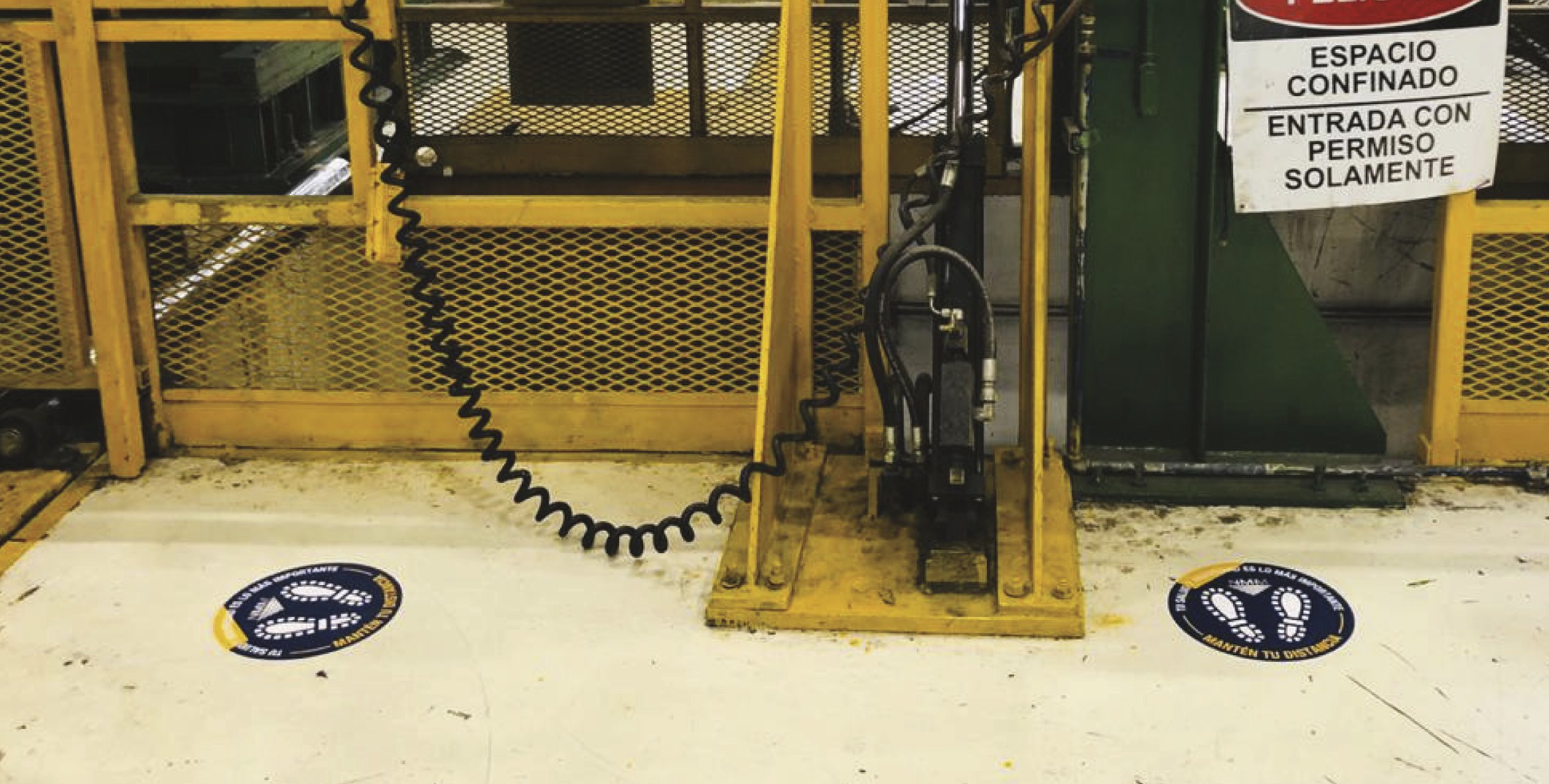Industrial safety markers on the floor indicating social distancing in Spanish, with yellow safety barriers and machinery in the background.