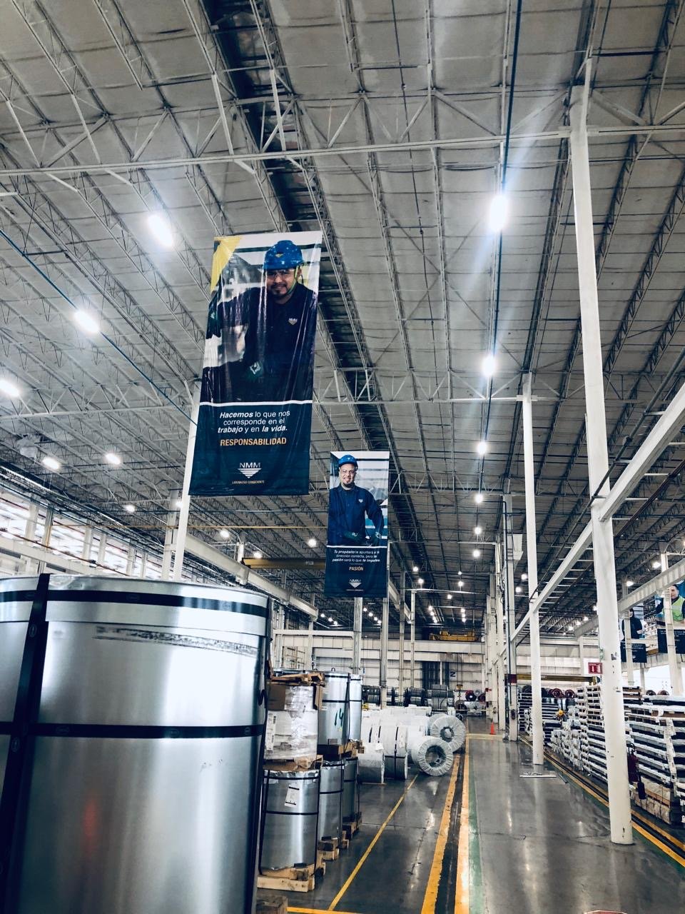 Inside a large warehouse, tall banners featuring workers in safety helmets and uniforms hang from the ceiling. The storage area is filled with large rolls of metal sheets and other materials stacked on pallets and shelves.