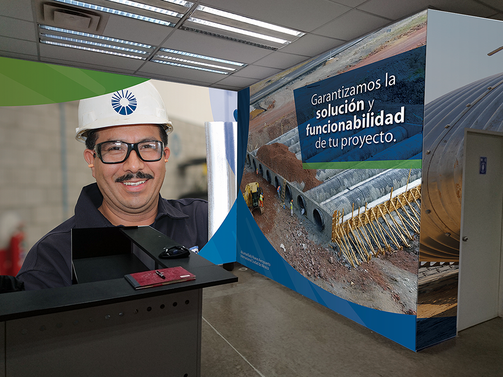 Office reception desk with a man wearing glasses, a helmet, and a dark shirt, smiling. Behind him is a large banner in Spanish with images of construction pipes, construction site, and the text 'Garantizamos la solución y funcionalidad de tu proyecto