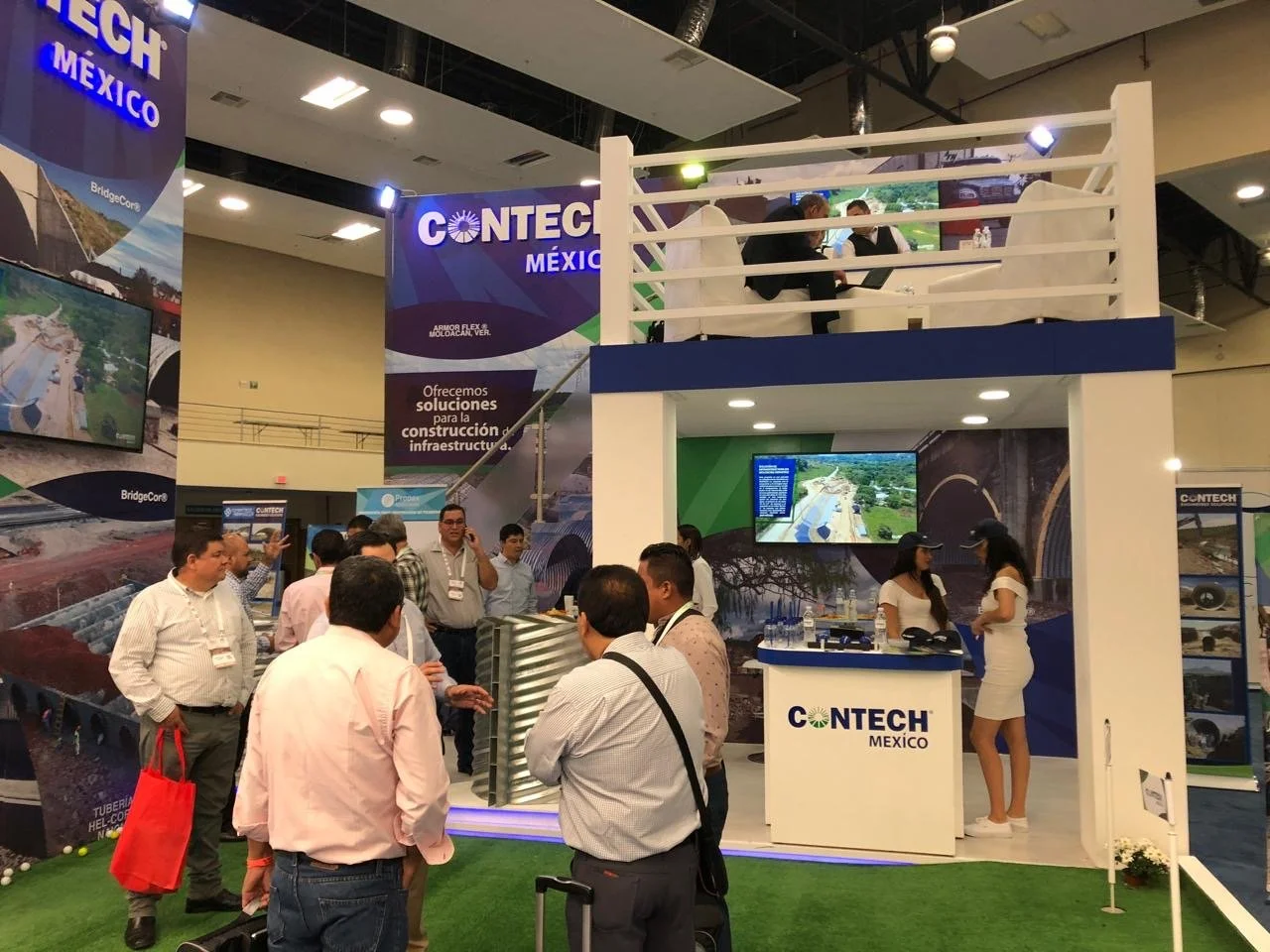 Exhibition booth of CONTECH México at a trade show, featuring displays of infrastructure projects, a large screen showing construction images, and two women in white dresses talking, with several attendees engaging in conversations.