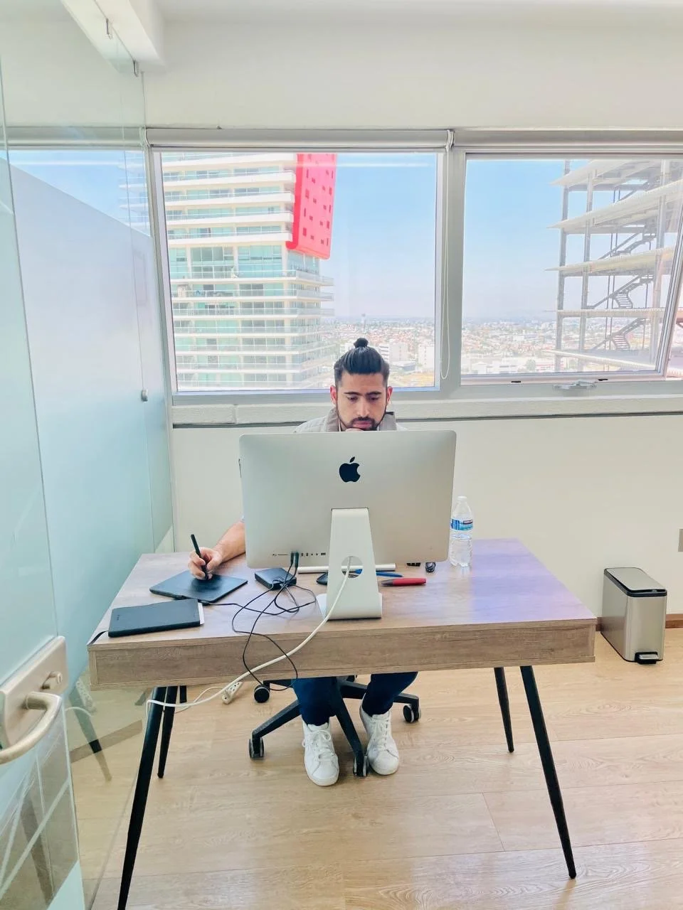 A man with a top bun hairstyle working at a desk with an iMac computer, a water bottle, a phone, and notebook, in a modern office with city view through large windows.
