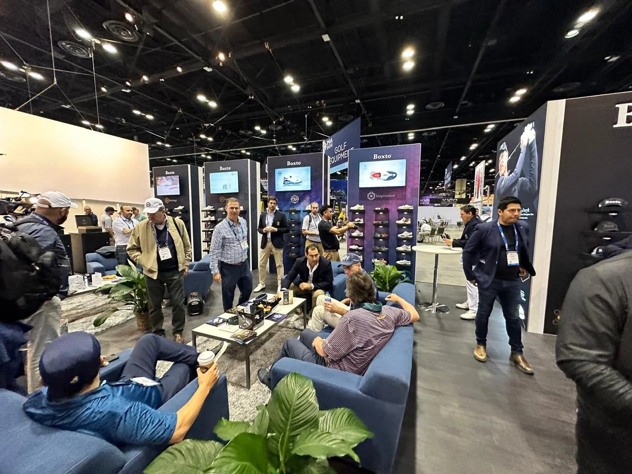 People attending a trade show or exhibition in a large indoor venue, with booths displaying golf shoes and equipment, some seated and others mingling or browsing