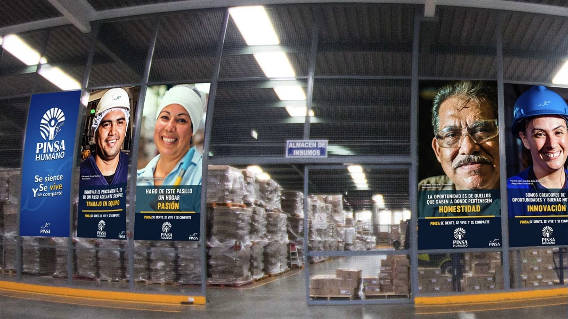 Inside a warehouse with promotional posters on the wall, featuring diverse workers and slogans about teamwork, honesty, and innovation. The warehouse has shelves with stacked boxes and supplies.