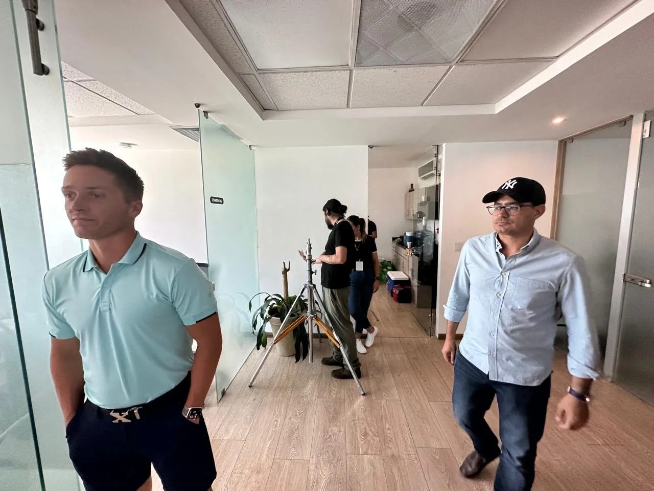 Office hallway with two men walking, one in a light blue polo shirt and dark pants, the other in a light blue shirt, jeans, and a black cap, with a water cooler and people working on a film set in the background.