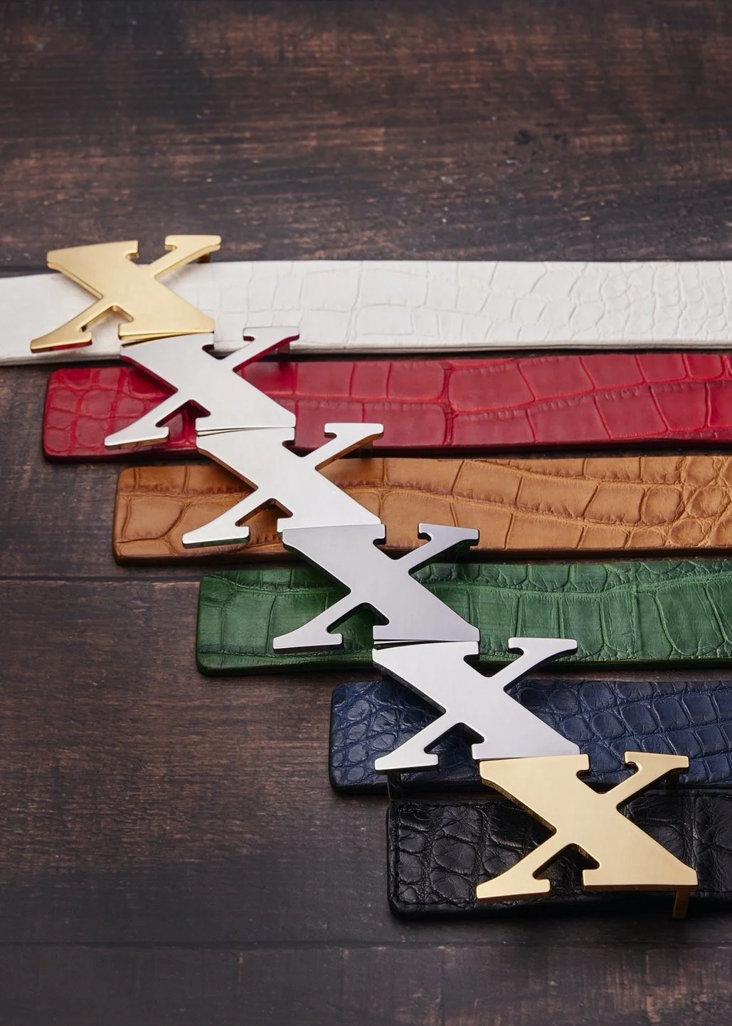 Collection of five leather belts in black, green, blue, brown, red, and white, each with a large metal buckle shaped like a zigzag or stylized 'X,' laid out on a wooden surface.
