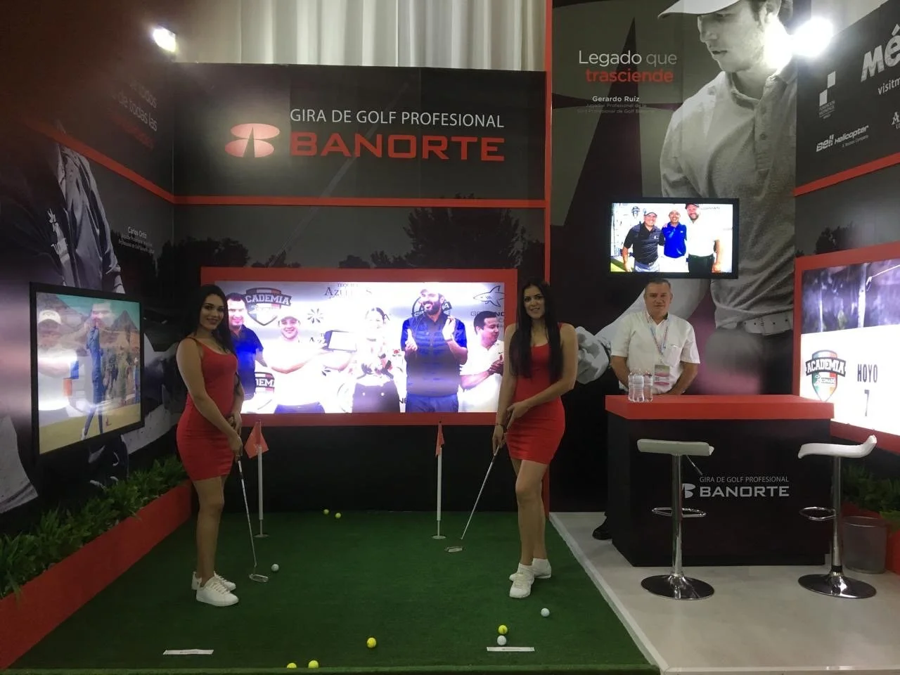 A golf-themed promotional booth for BANORTE, featuring two women in red dresses holding golf clubs, a man in white behind a counter, three TV screens displaying golf images, and a backdrop with a golfer and promotional text.