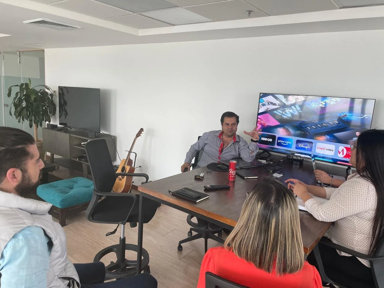 A group of five people in a meeting room, with one person presenting while seated at a large table. The presenter is gesturing, with a television displaying streaming service logos behind him. A guitar is leaning against a chair, and a large TV is on