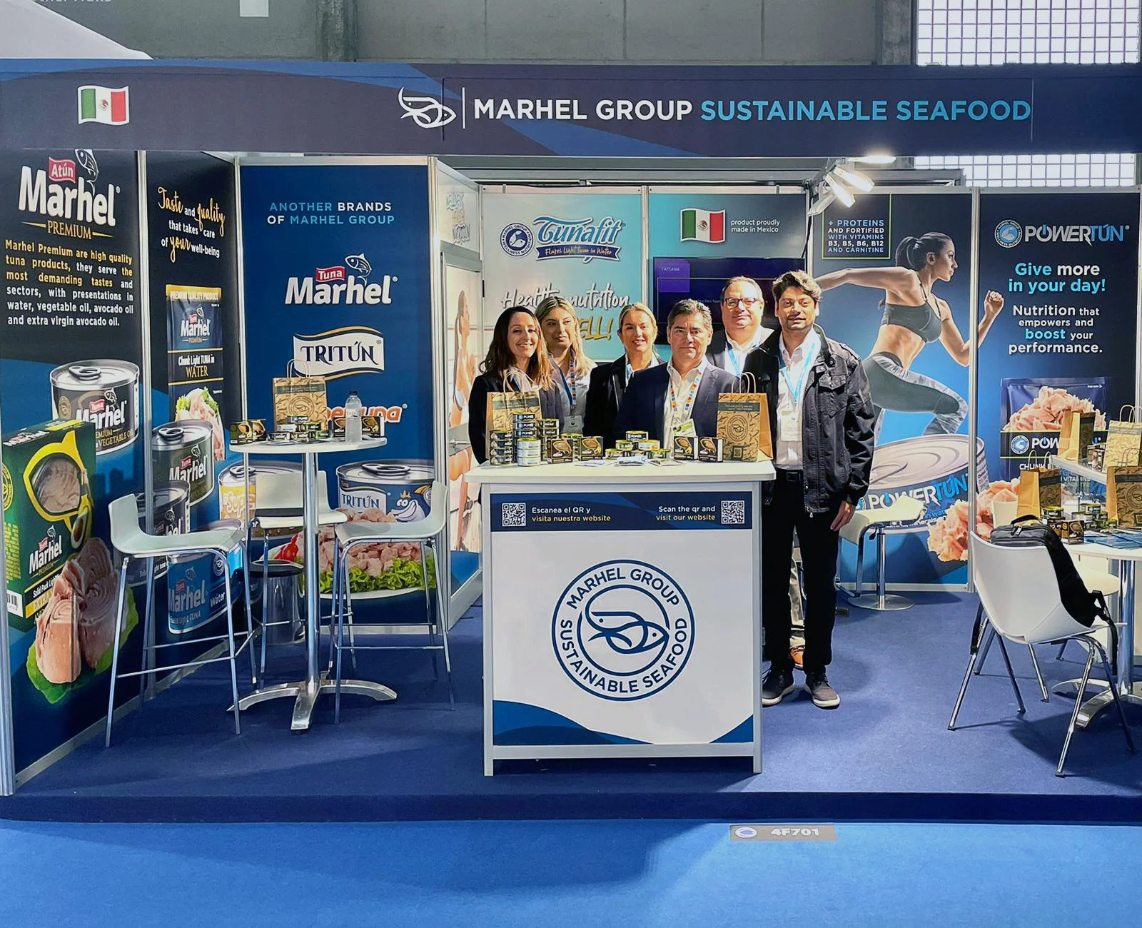 Five people standing behind a booth display at a trade show for Marhel Group's sustainable seafood products. The booth features promotional posters, product samples, and a logo on the front desk.