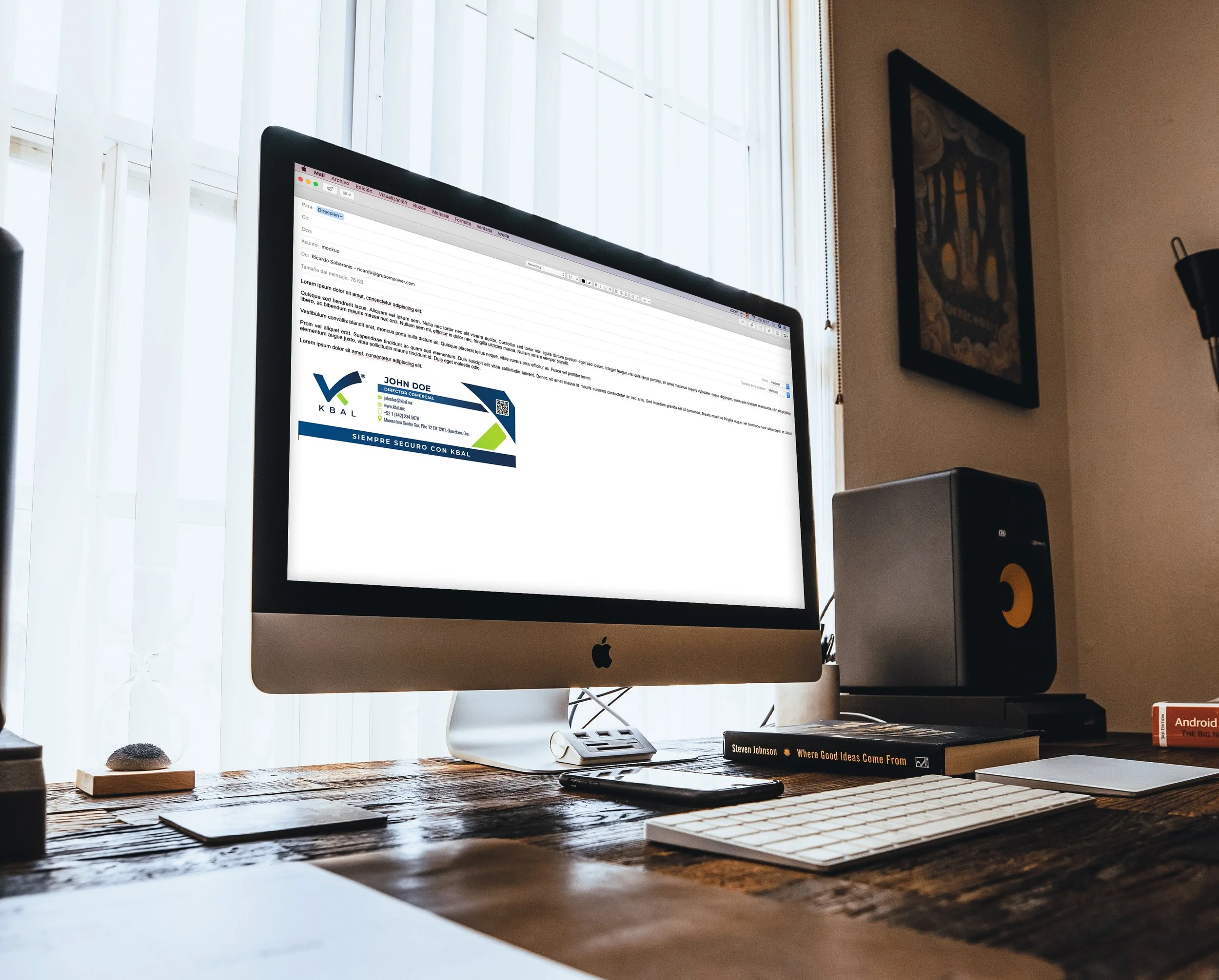 An Apple iMac computer on a wooden desk displaying an email with a business card attached. The desk has a book, a smartphone, a wireless keyboard, and a wireless mouse. There is a speaker and a book titled 'Where Good Ideas Come From' on the right si