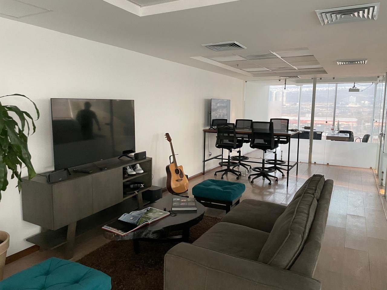 Modern office lounge and work area with a flat-screen TV, guitar, armchair, and high-top tables with chairs, large windows with city view