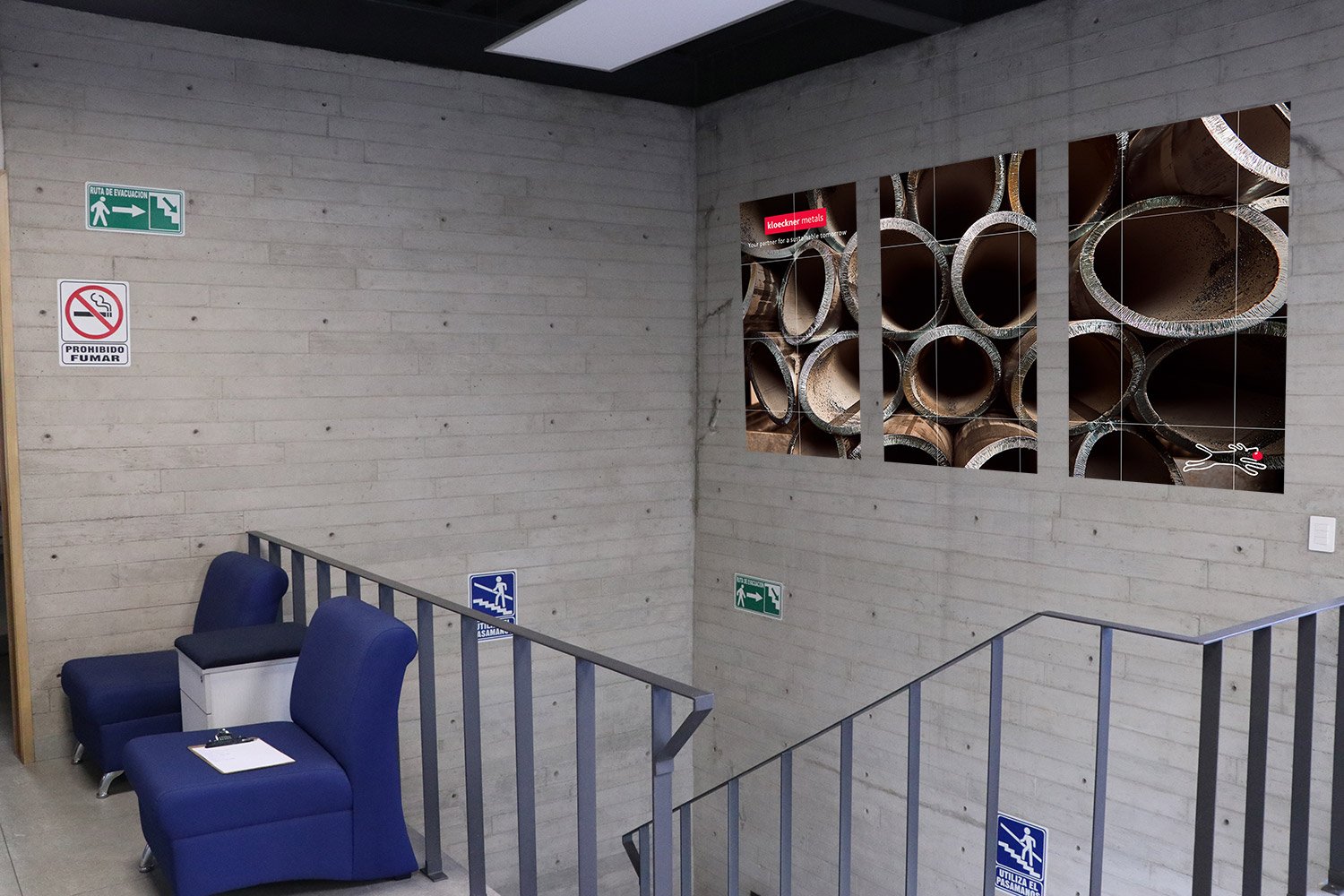 Indoor staircase area with concrete walls, blue chairs, and safety signs including emergency exit and no smoking. Artistic large photograph of industrial pipes on the wall.