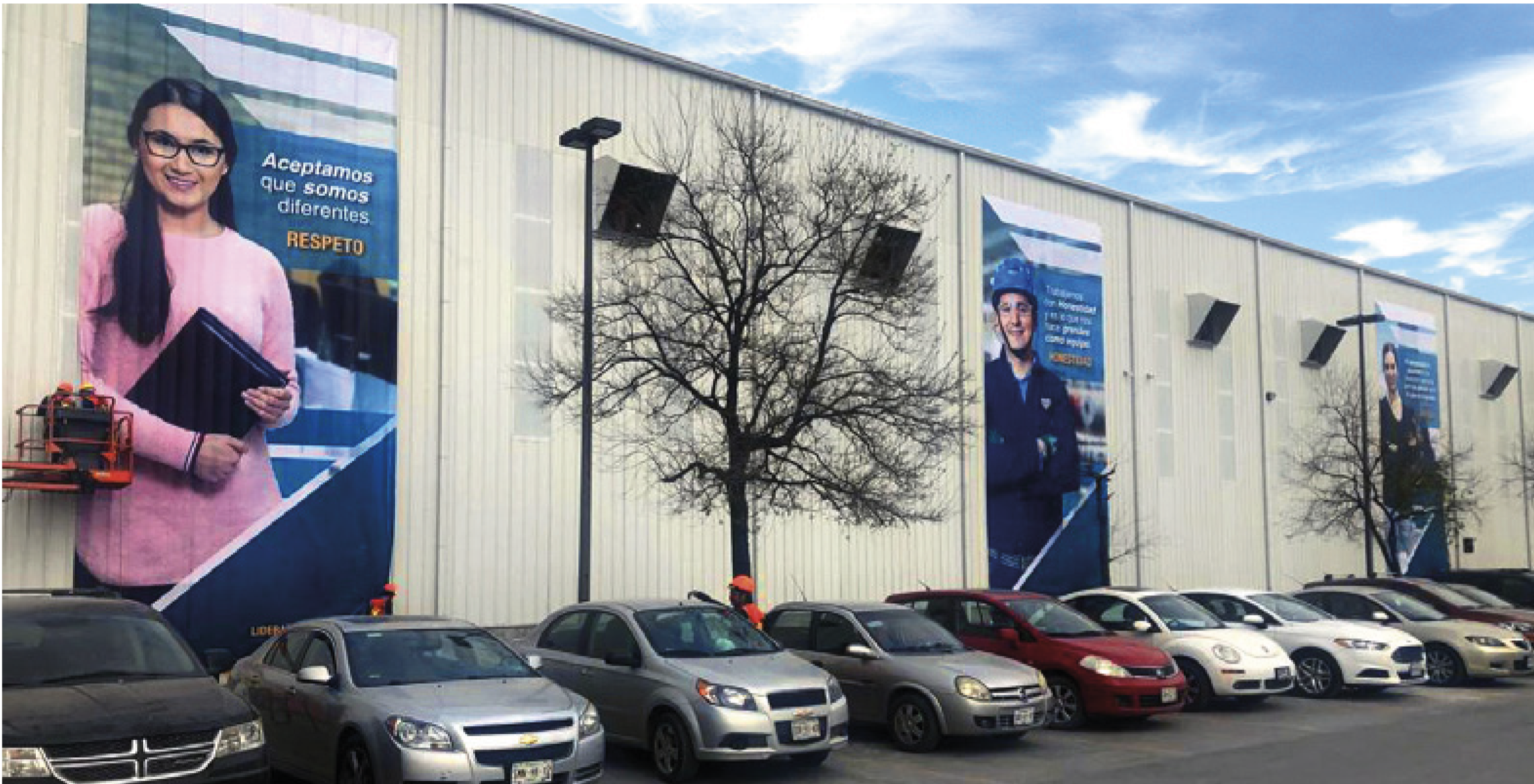 Large building exterior with three prominent posters on the wall, each featuring people and Spanish text, including a woman holding folders and a man with crossed arms in work attire, with a parking lot in front containing various cars and two leafle