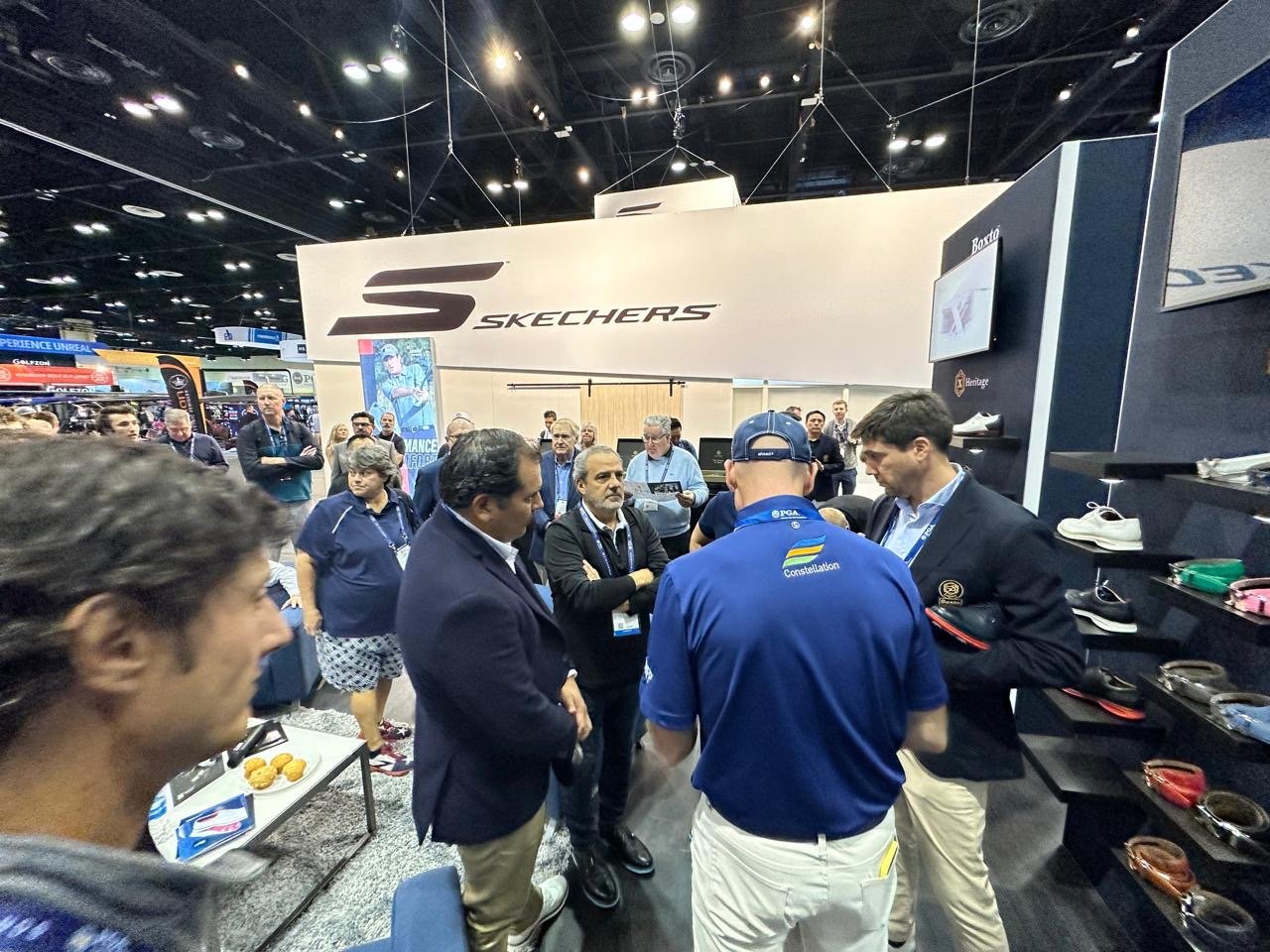 Group of men conversing at the Skechers booth during a trade show or exhibition, with shoes displayed on shelves and a large Skechers sign on the backdrop.