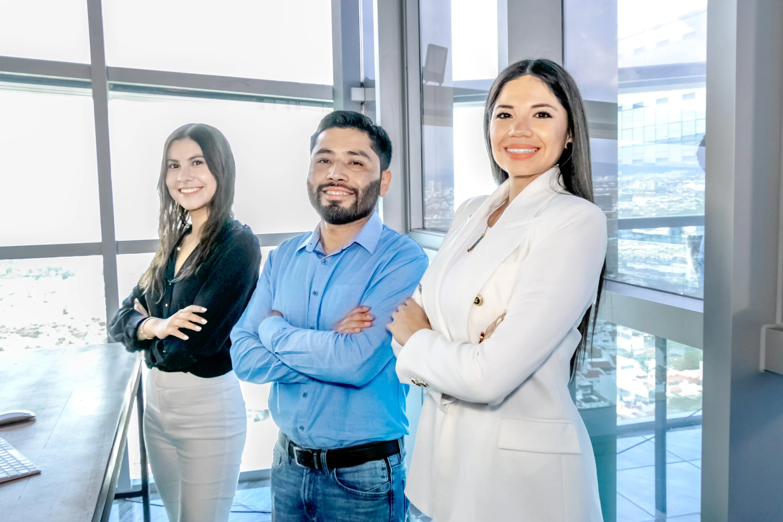 Three diverse professionals standing with arms crossed in a modern office with large windows and city view, smiling at the camera.
