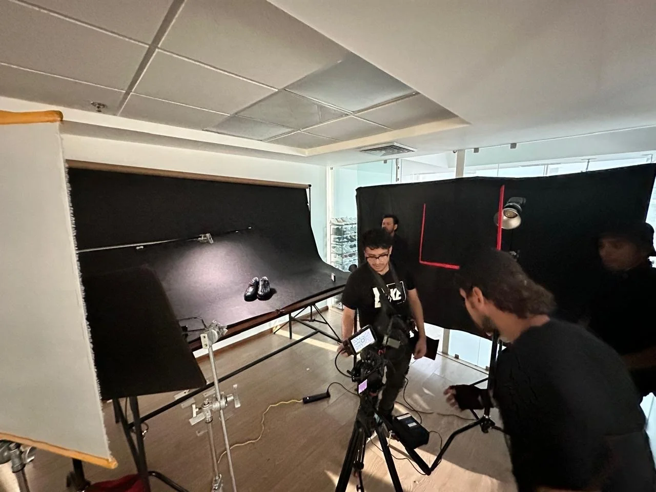 A behind-the-scenes look at a photo or video shoot with a black backdrop, lighting equipment, a camera on a tripod, and a few crew members preparing.