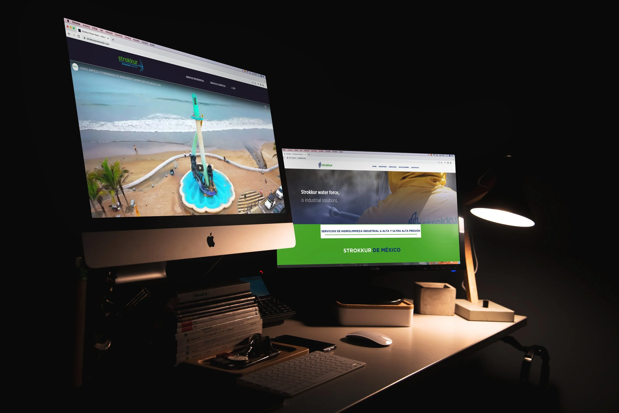 A computer workstation with two monitors displaying websites, a desk lamp, mouse, keyboard, and office supplies in a dimly lit room.