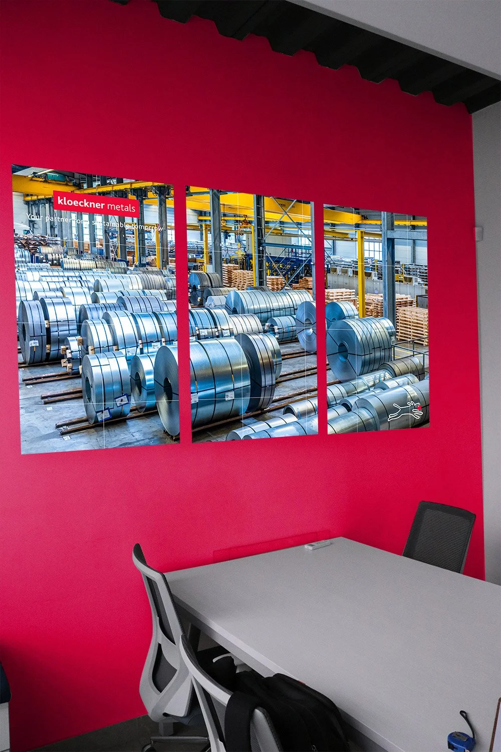 Picture of a large, industrial warehouse filled with rows of shiny steel coils and rolls stored on the floor, viewed through three glass panels on a bright red wall in an office setting.